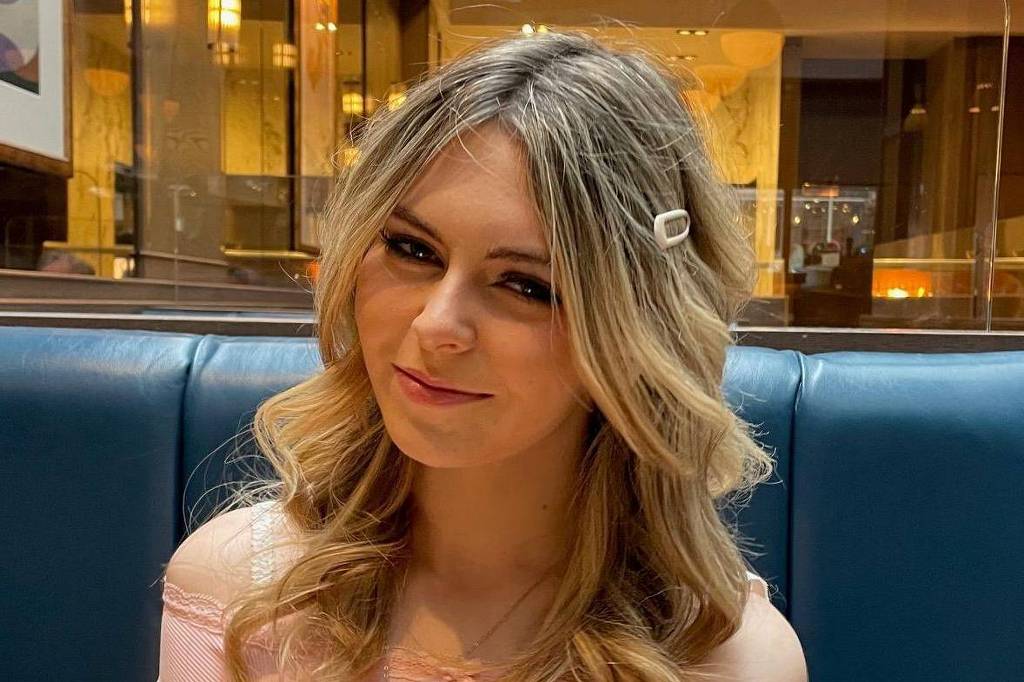 Mulher jovem com cabelos loiros ondulados presos parcialmente por presilha branca, sentada em banco azul em ambiente interno com iluminação amarelada e paredes de vidro ao fundo.