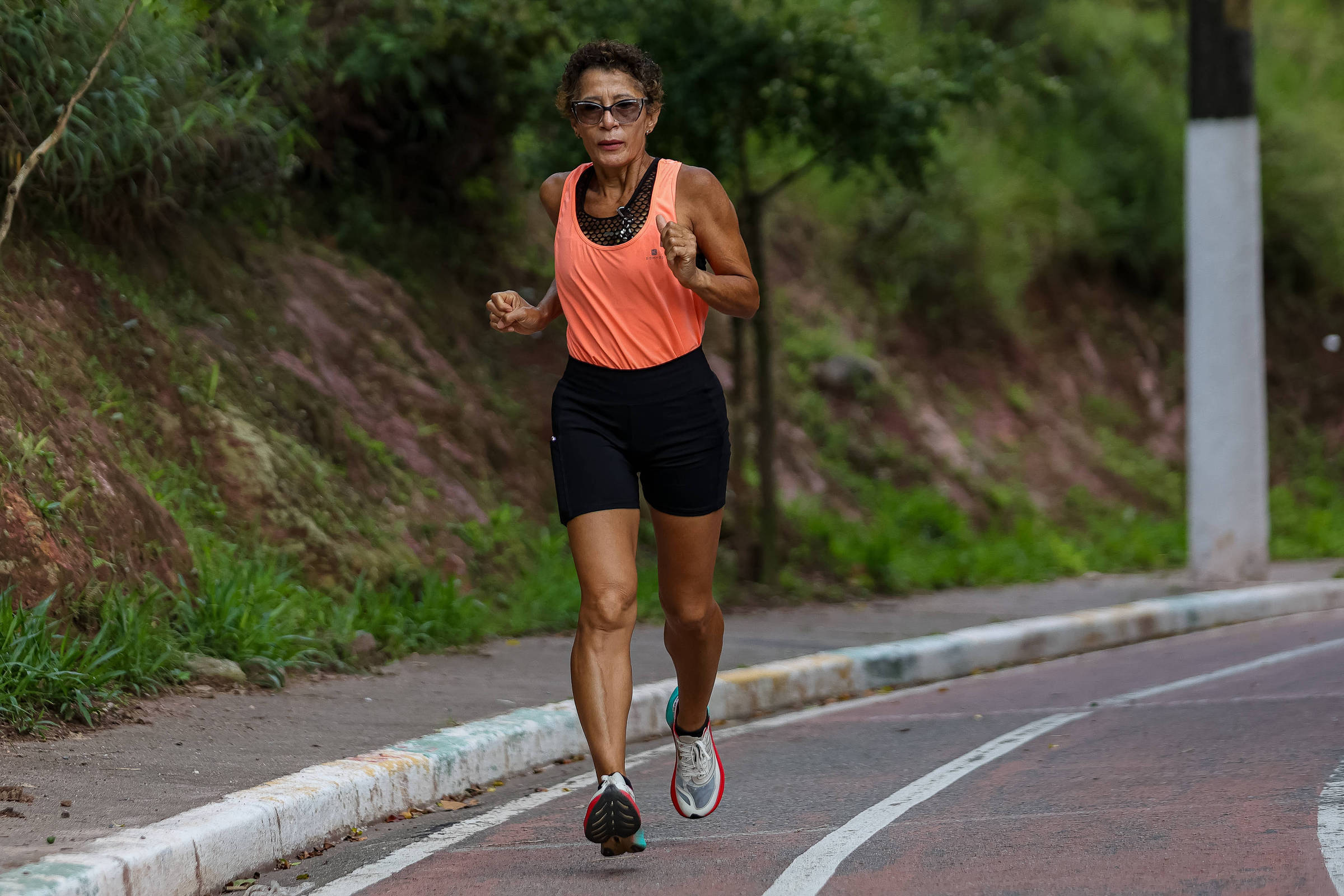 Mulher adulta corre em pista de corrida ao ar livre, vestindo regata laranja e shorts pretos. Vegetação e poste aparecem ao fundo.