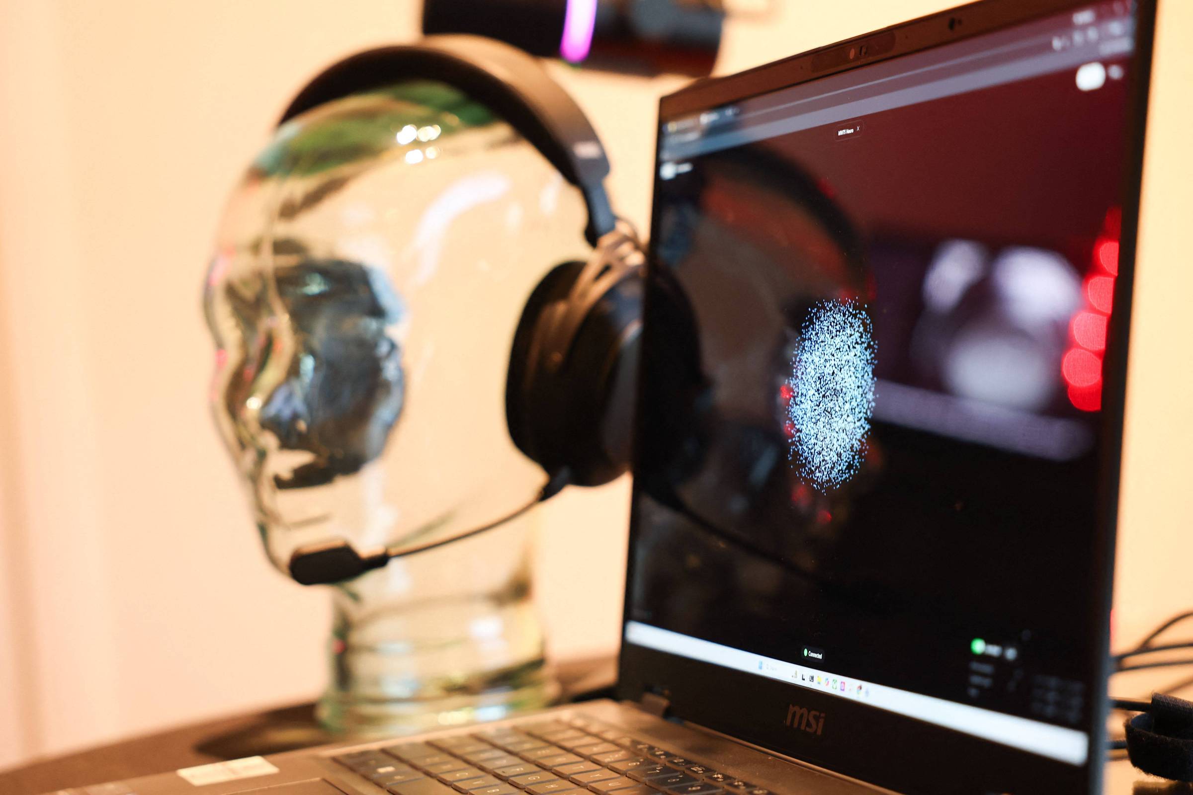 Cabeça de vidro transparente com fone de ouvido preto apoiada sobre mesa ao lado de laptop aberto. Tela do laptop mostra imagem pixelada de rosto humano.