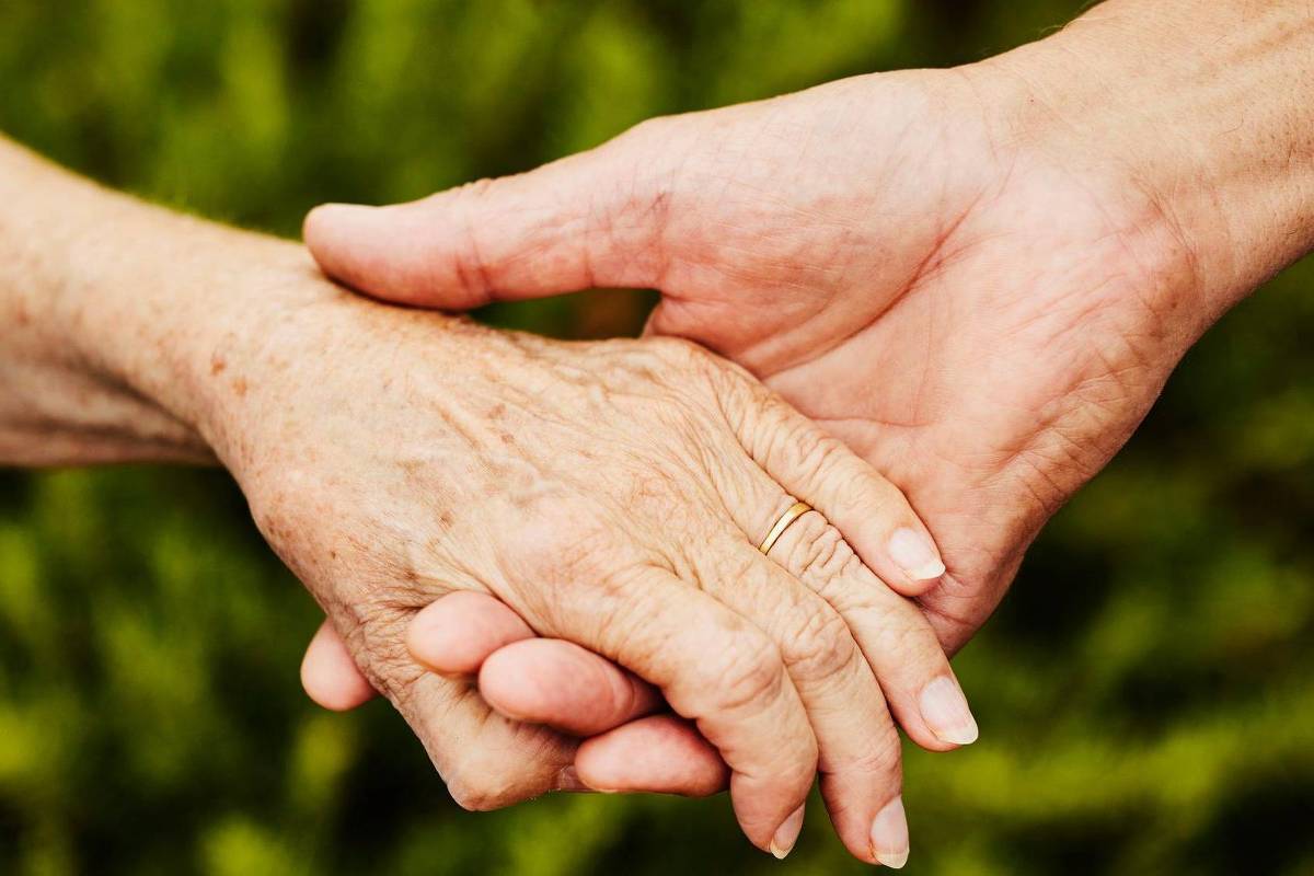 Mãos de duas pessoas idosas entrelaçadas, uma com aliança dourada, sobre fundo desfocado de vegetação verde.