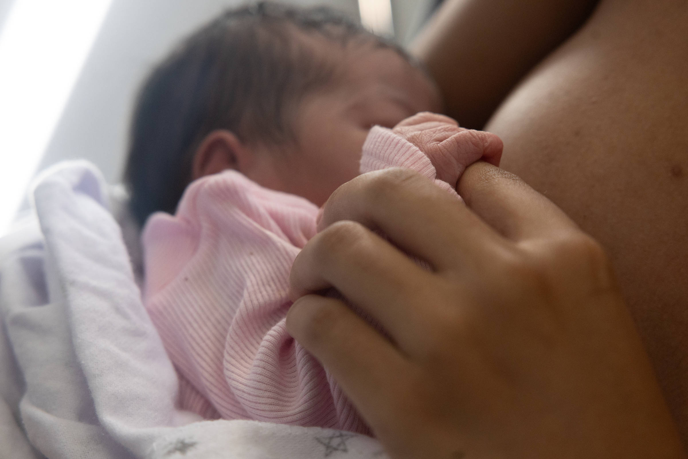 A imagem mostra um bebê sendo amamentado. O bebê está vestido com uma roupa rosa e é segurado por uma mão que parece ser de um adulto. A mão do adulto está segurando delicadamente a mão do bebê. O fundo é desfocado, mas parece haver uma fonte de luz suave.