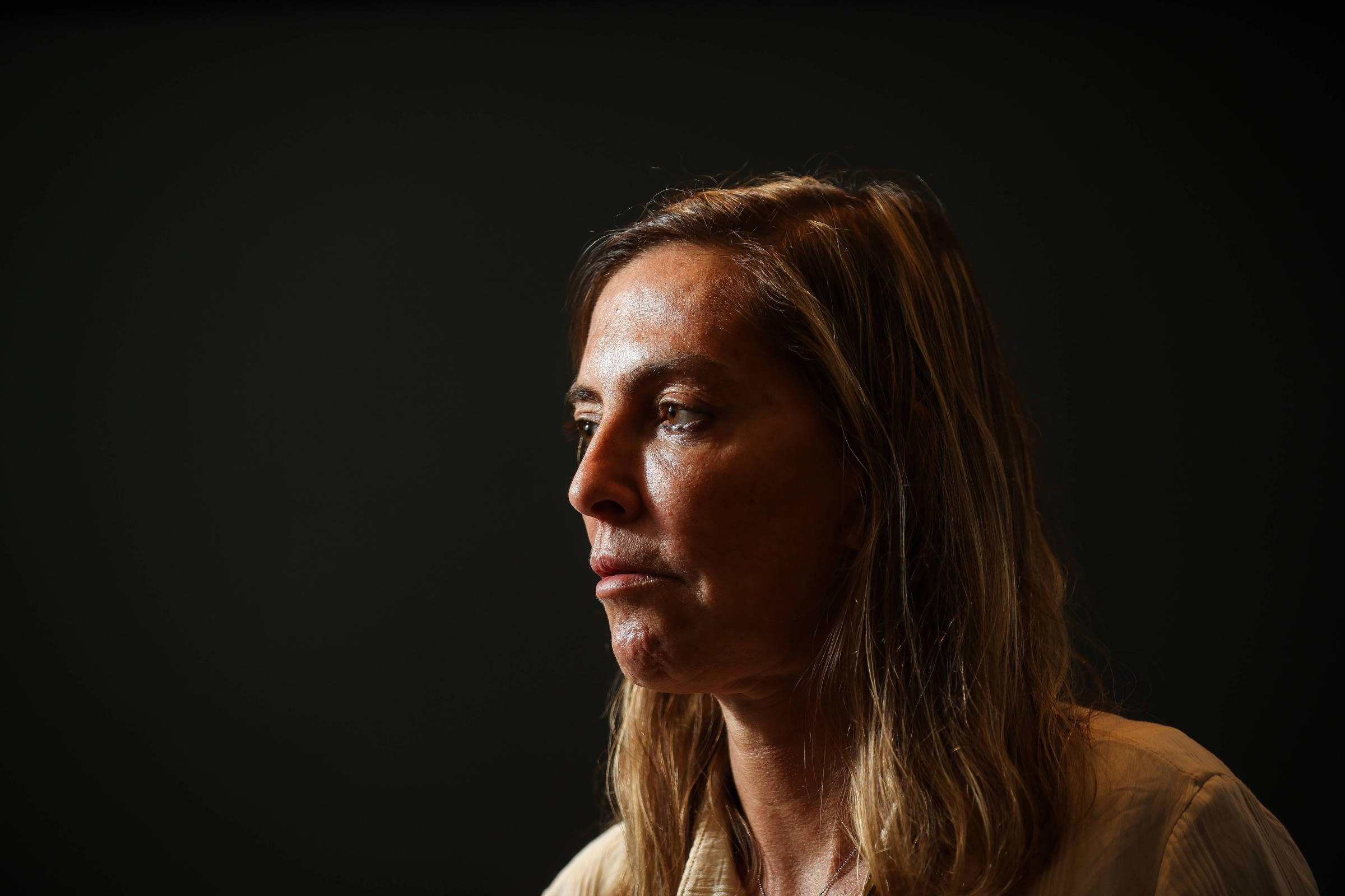 Mulher de cabelos castanhos claros e lisos, olhando para a esquerda, com expressão neutra. Fundo preto uniforme destaca seu rosto iluminado de perfil.