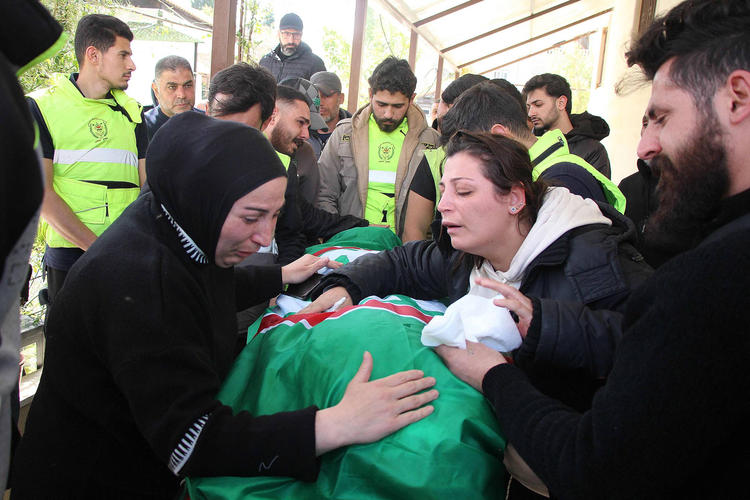 Grupo de pessoas em ambiente externo coberto, algumas vestindo coletes amarelos, rodeiam um corpo coberto por uma bandeira verde, vermelha e branca. Duas mulheres na frente demonstram tristeza, uma delas segura um lenço branco.