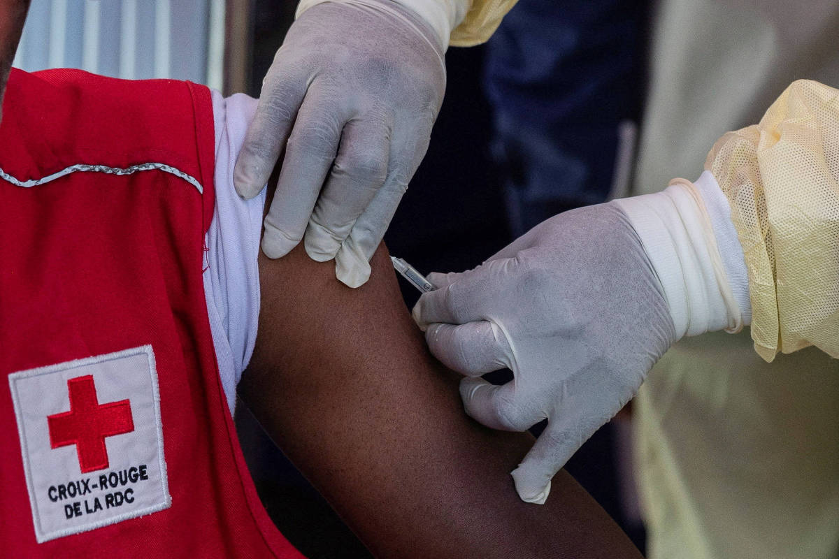 Profissional com luvas e jaleco amarelo aplica vacina no braço de pessoa vestindo colete vermelho da Cruz Vermelha da República Democrática do Congo.