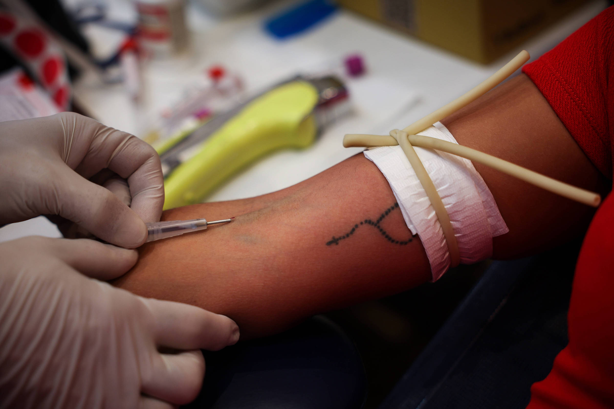 Braço de mulher com elástico fazendo aperto, enquanto profissional de saúde coloca agulha no antebraço para retirar sangue