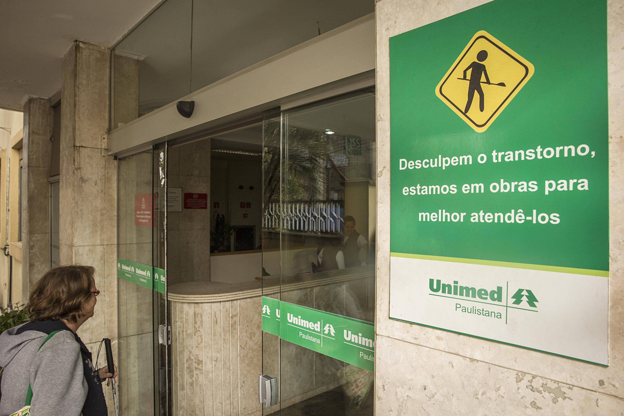 Placa verde na parede ao lado da entrada de vidro da Unimed Paulistana informa sobre obras em andamento para melhor atendimento. Mulher observa a entrada do prédio.