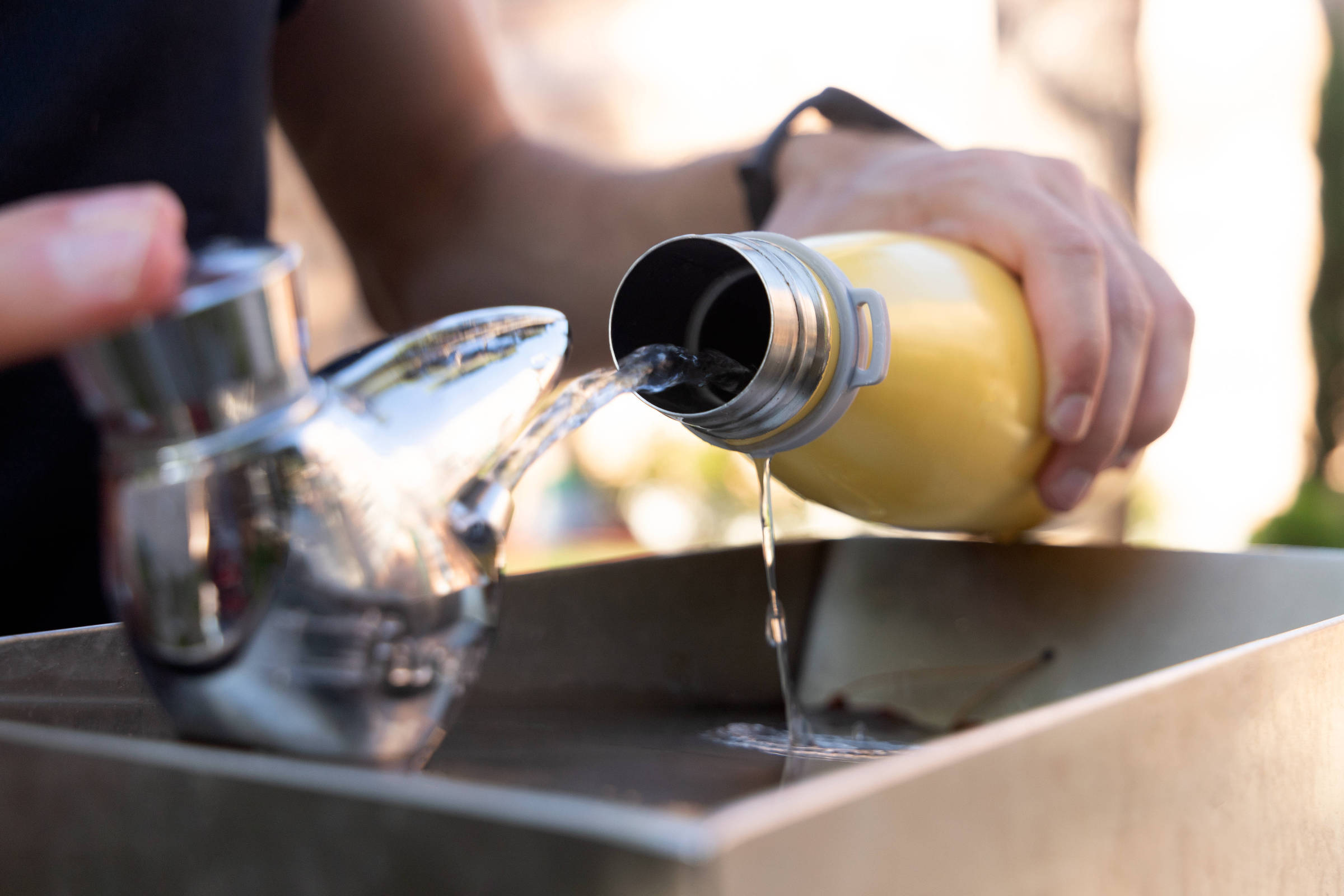 Mãos seguram uma garrafa amarela e despejam líquido em um bule metálico prateado sobre uma superfície retangular. Fundo desfocado com luz natural.