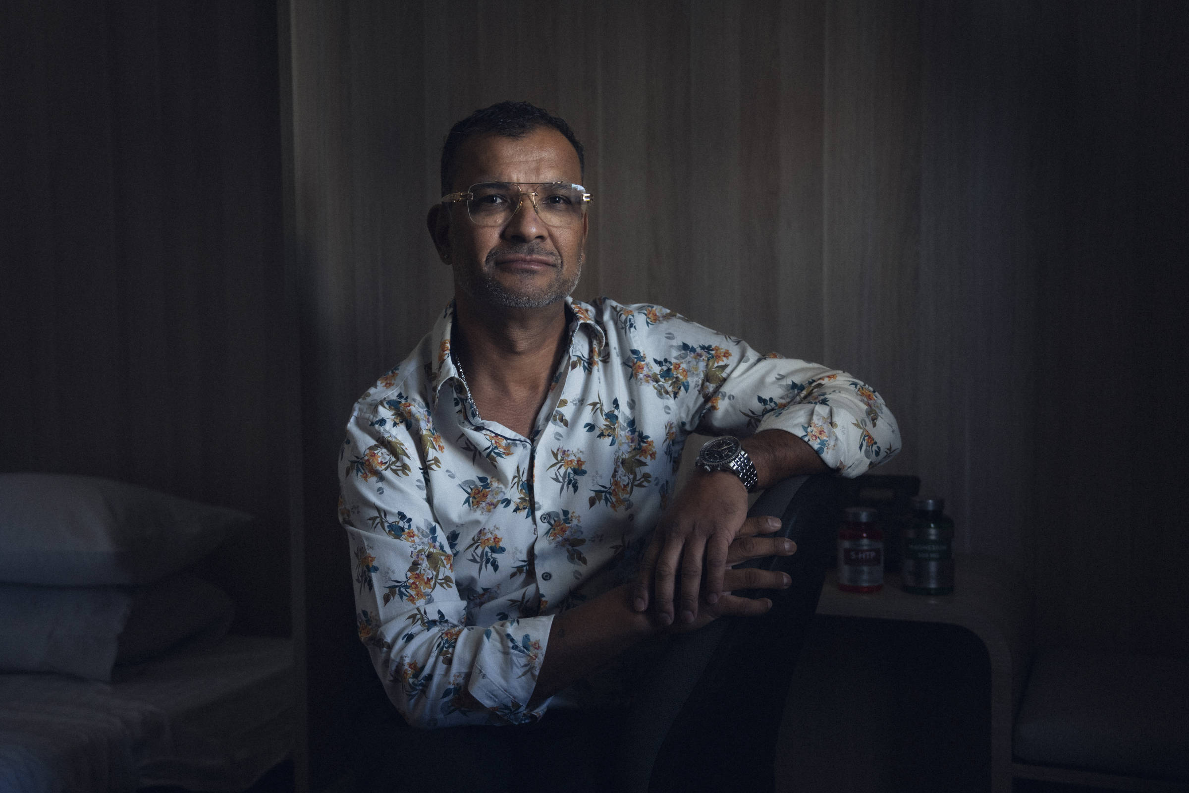 Homem de meia-idade com camisa branca estampada com flores, usando óculos, sentado com um braço apoiado no joelho em ambiente interno com parede de madeira ao fundo. Ao lado, mesa pequena com frascos e cama com travesseiros empilhados.