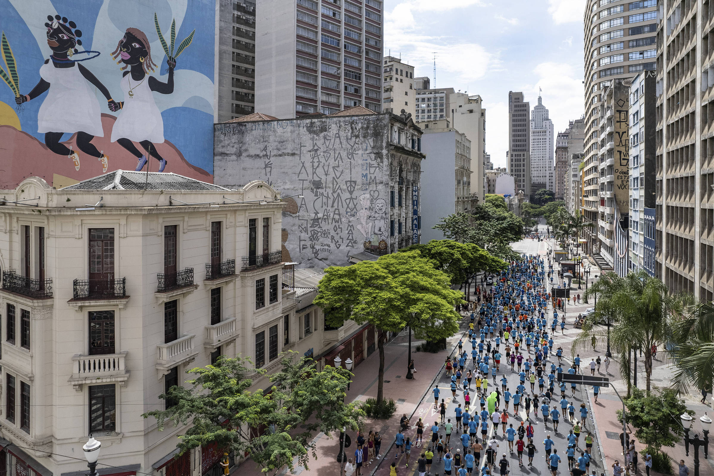Centenas de pessoas caminham pela Avenida São João, em São Paulo, em dia claro. À esquerda, prédio antigo com sacadas e mural colorido com figuras femininas negras segurando plantas. Ao fundo, prédios altos e céu parcialmente nublado.