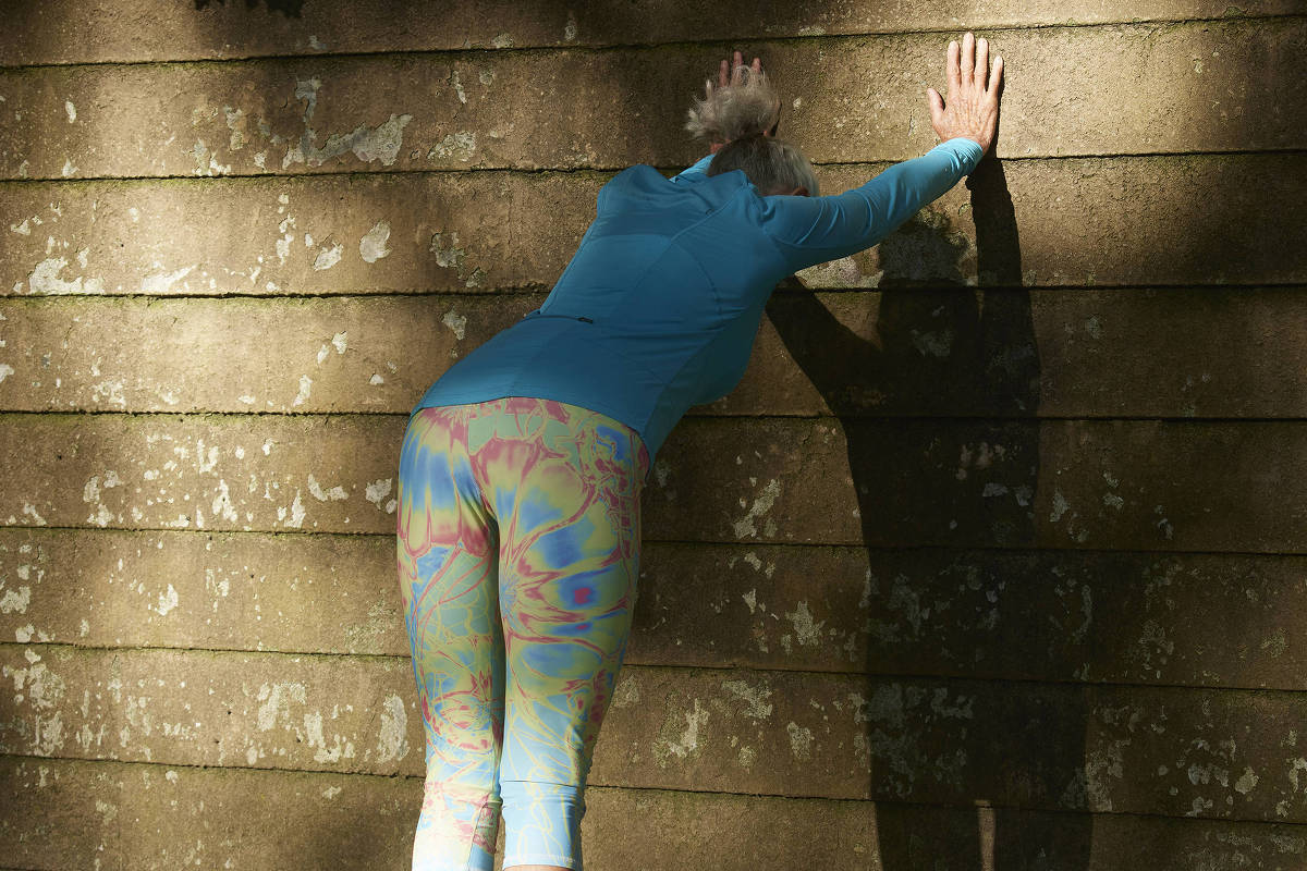 Pessoa vestindo camiseta azul e legging colorida encosta as mãos em parede de pedra para alongar. Sombra projetada na parede e chão com folhas secas.