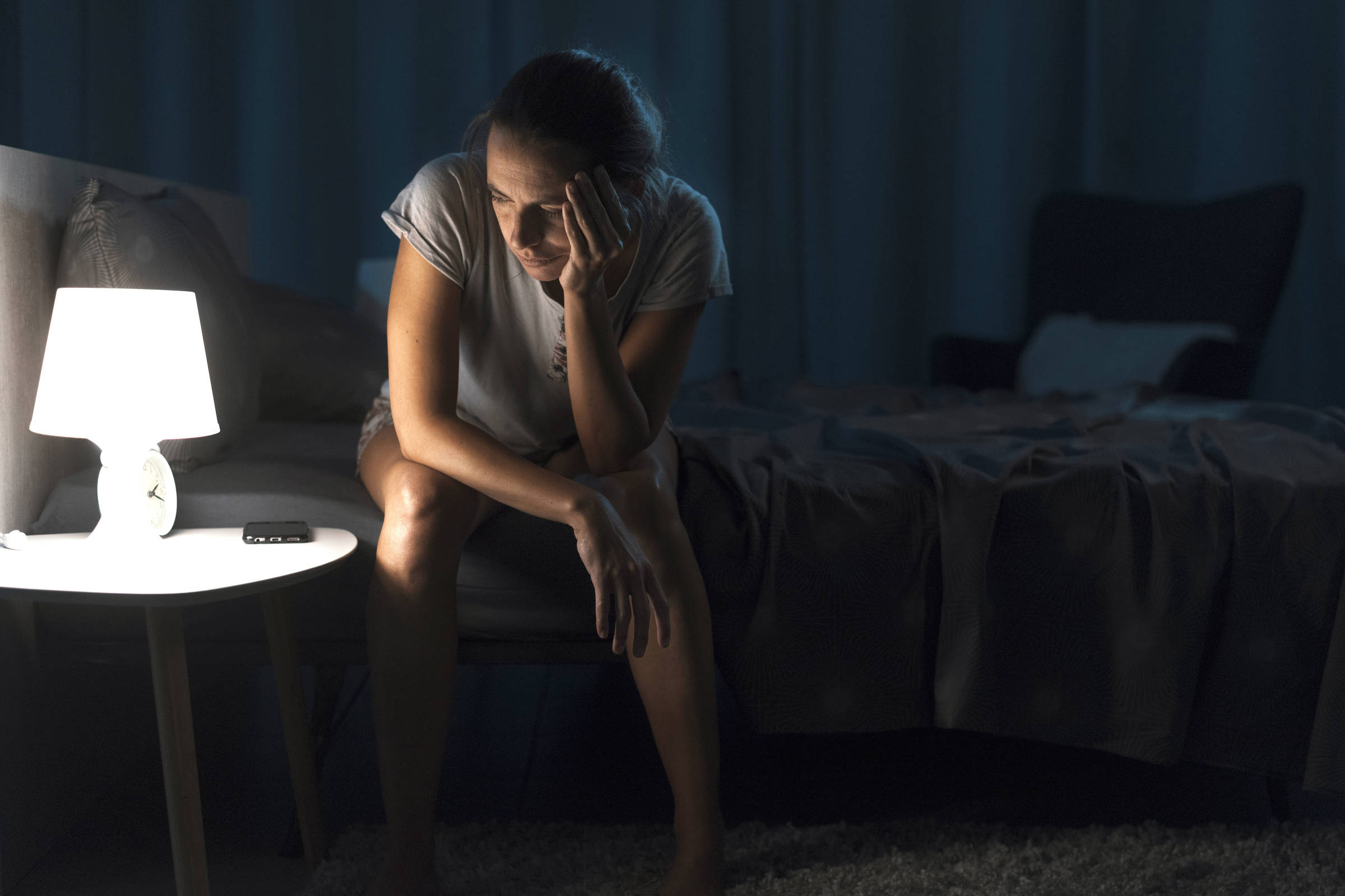 Mulher sentada na beira da cama em quarto escuro, iluminada por abajur branco sobre mesa lateral. Ela apoia a cabeça na mão, olhando para baixo, com controle remoto ao lado no móvel.