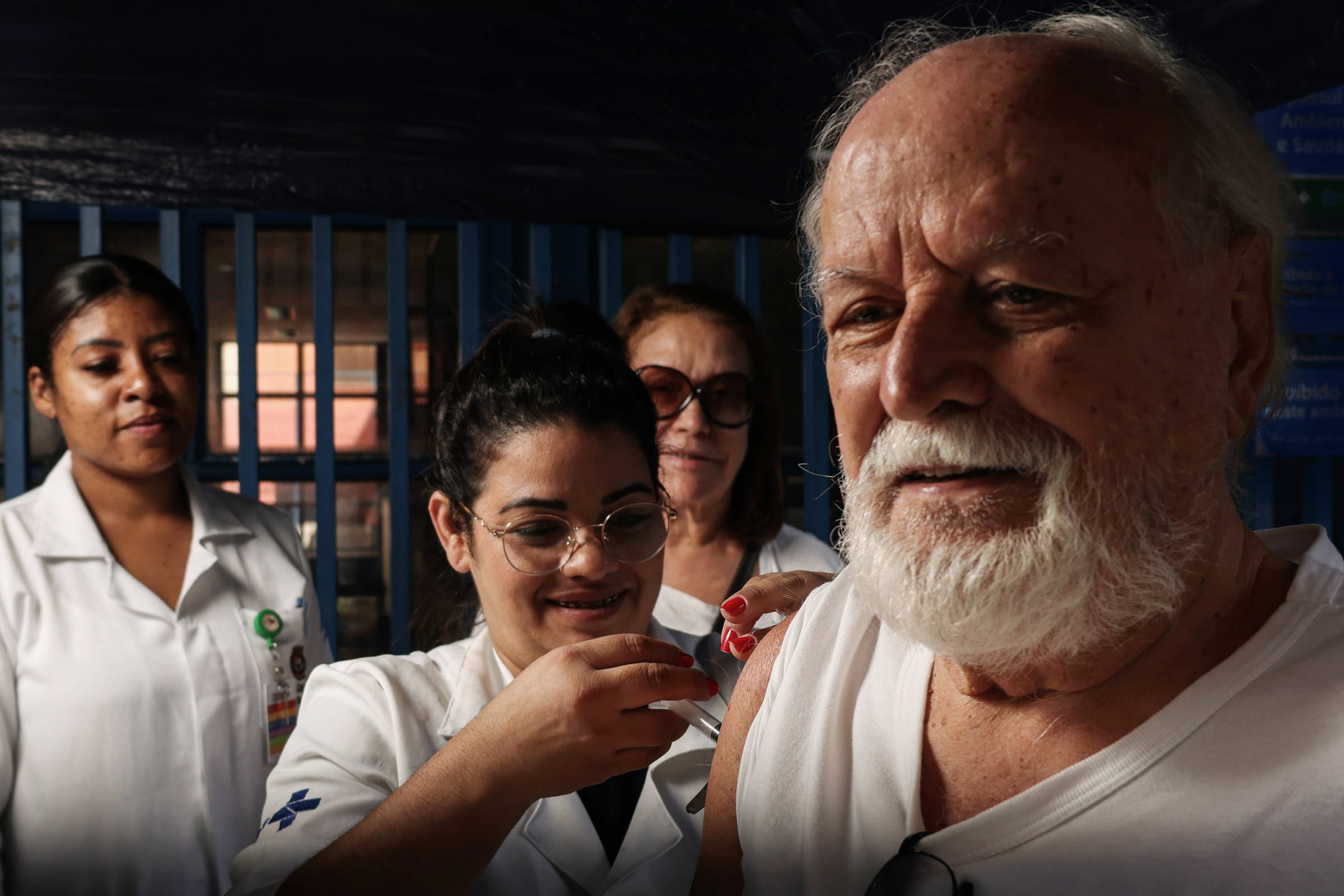 A imagem mostra um idoso com barba branca recebendo uma vacina. Ele está usando uma camiseta sem mangas e parece estar sorrindo. Ao fundo, duas profissionais de saúde, uma delas com óculos e a outra com um uniforme branco, observam e ajudam no processo de vacinação. O ambiente parece ser uma unidade de saúde, com uma grade azul ao fundo.