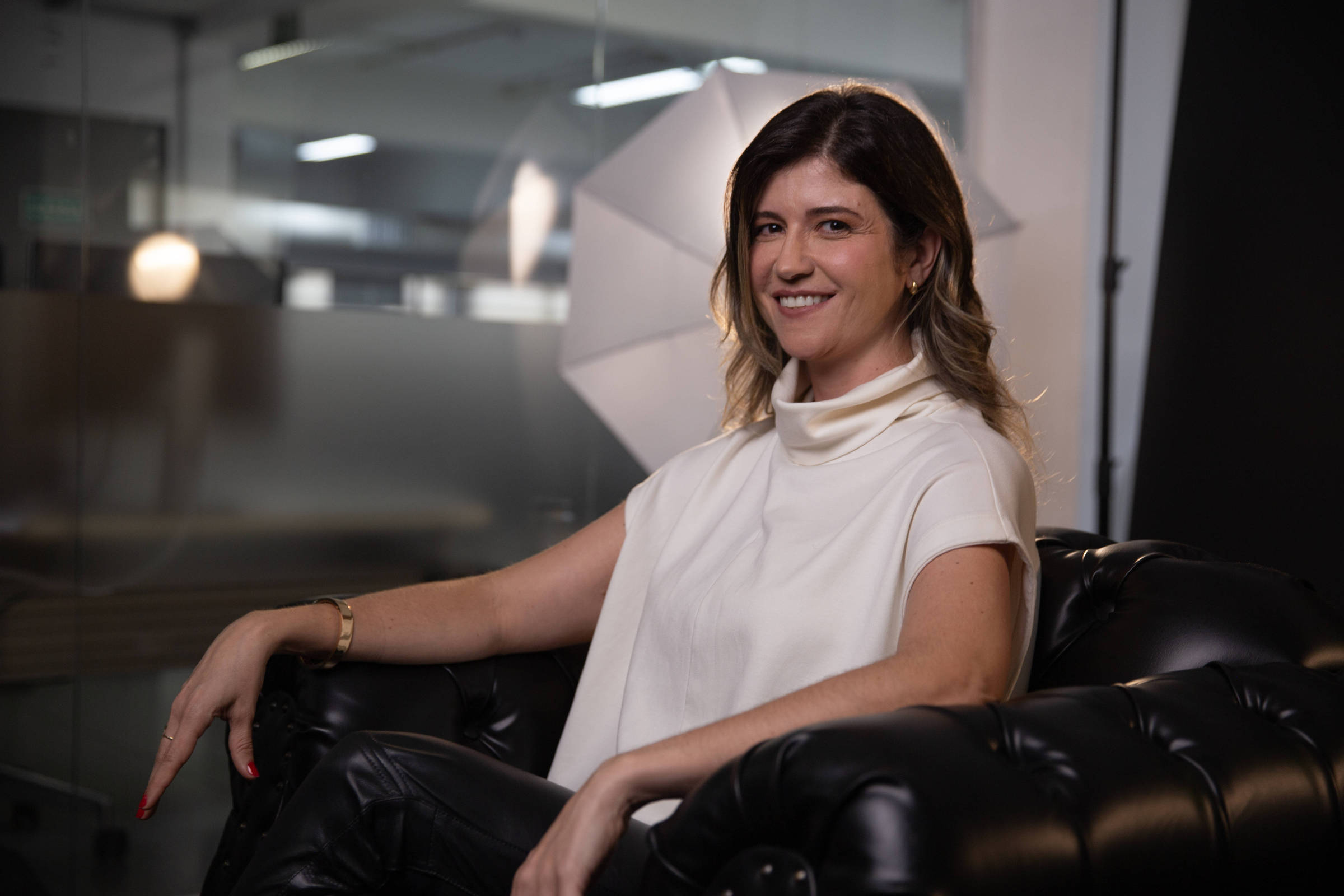 Foto de Aline Wolff sentada e sorrindo no estúdio da TV Folha. Ela é branca, tem cabelo liso, castanho escuro com pontas mais claras. Veste blusa branca e calça escura