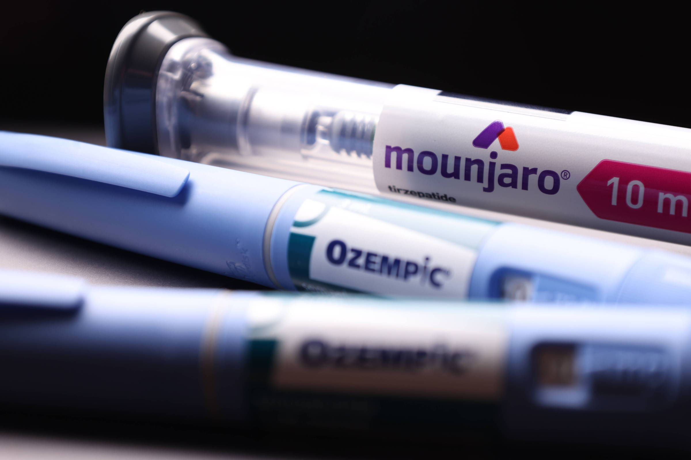 A imagem mostra três canetas de medicamento em primeiro plano. A caneta superior é transparente e contém o nome