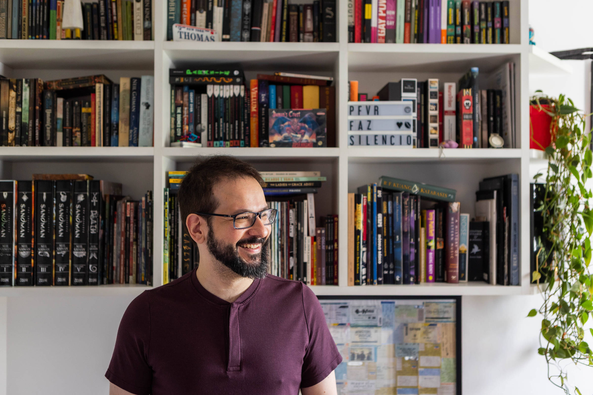 Homem de barba e óculos sorri olhando para o lado, em frente a uma estante branca cheia de livros e objetos. Na estante, há uma placa com a frase