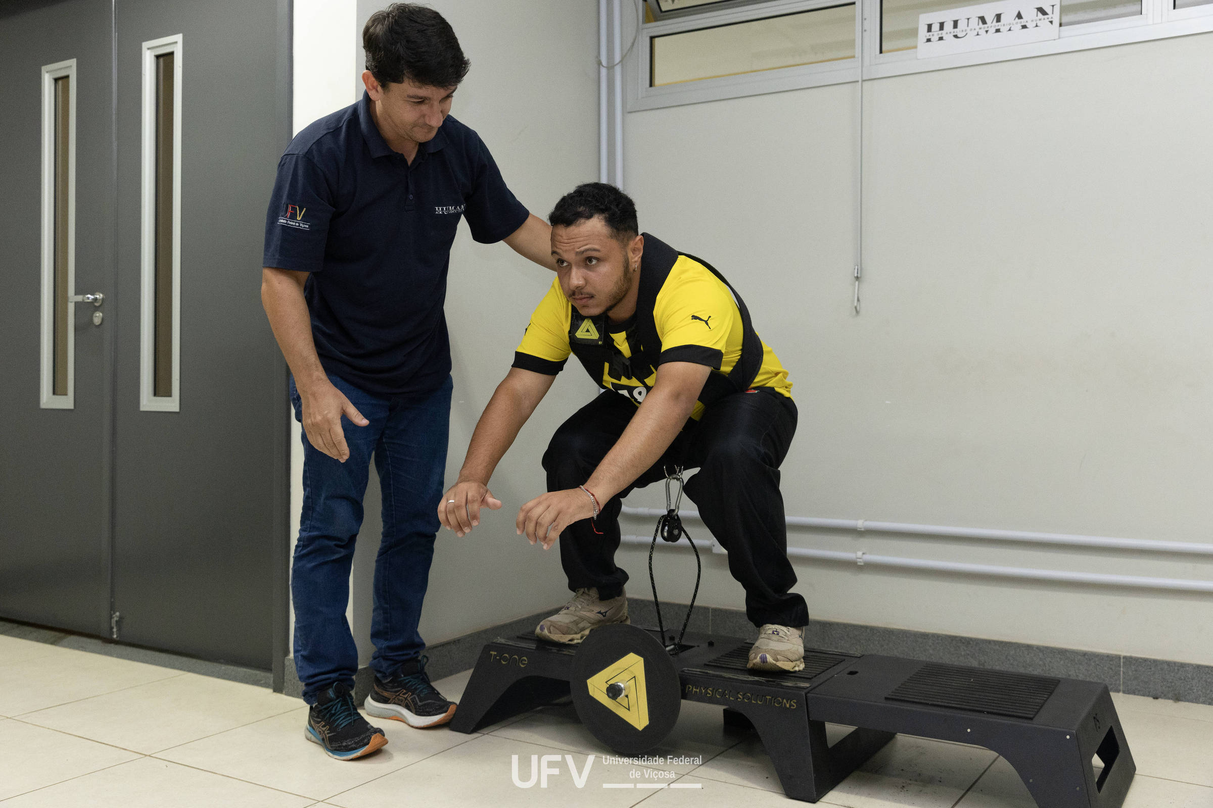 Homem agachado em equipamento de salto com sensores, acompanhado por outro homem que o orienta em ambiente interno.