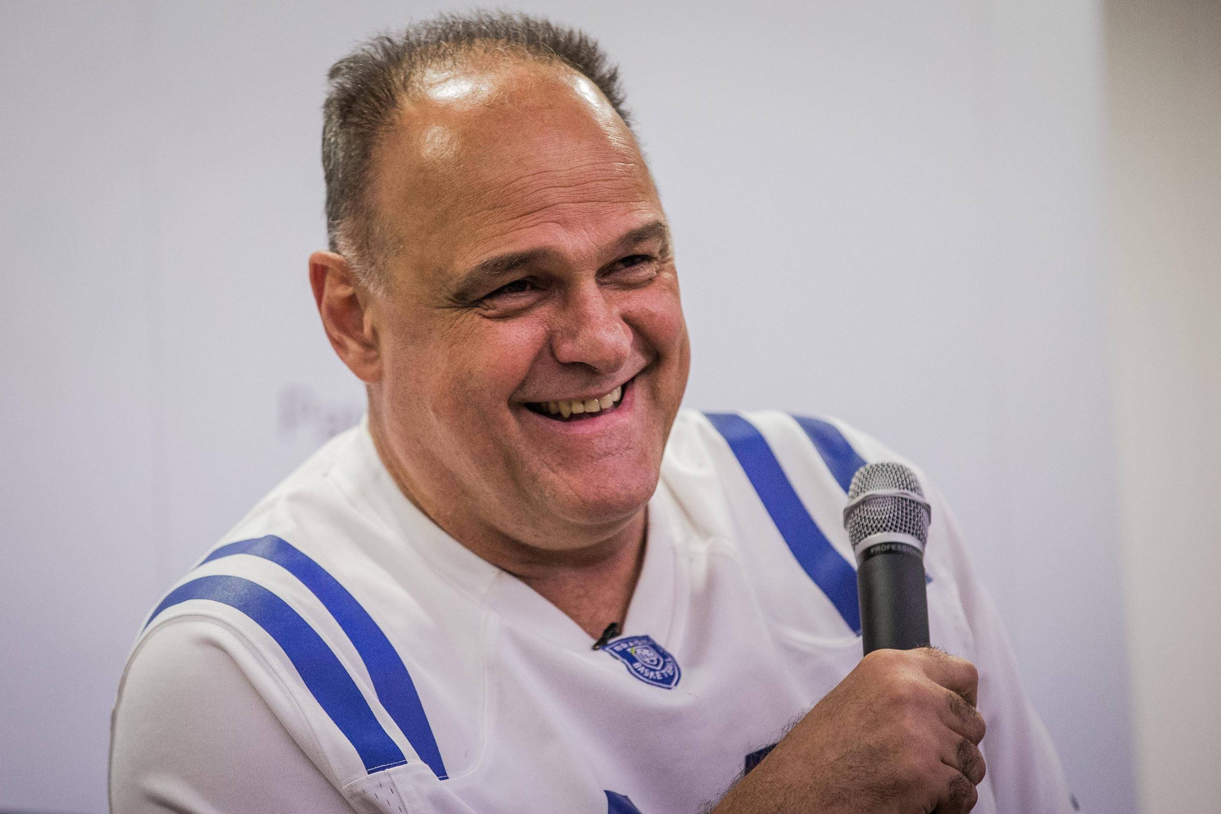 Homem de meia-idade com camiseta branca de mangas longas e detalhes azuis segura microfone com a mão direita. Ele sorri olhando levemente para a esquerda, em ambiente interno com fundo branco desfocado.