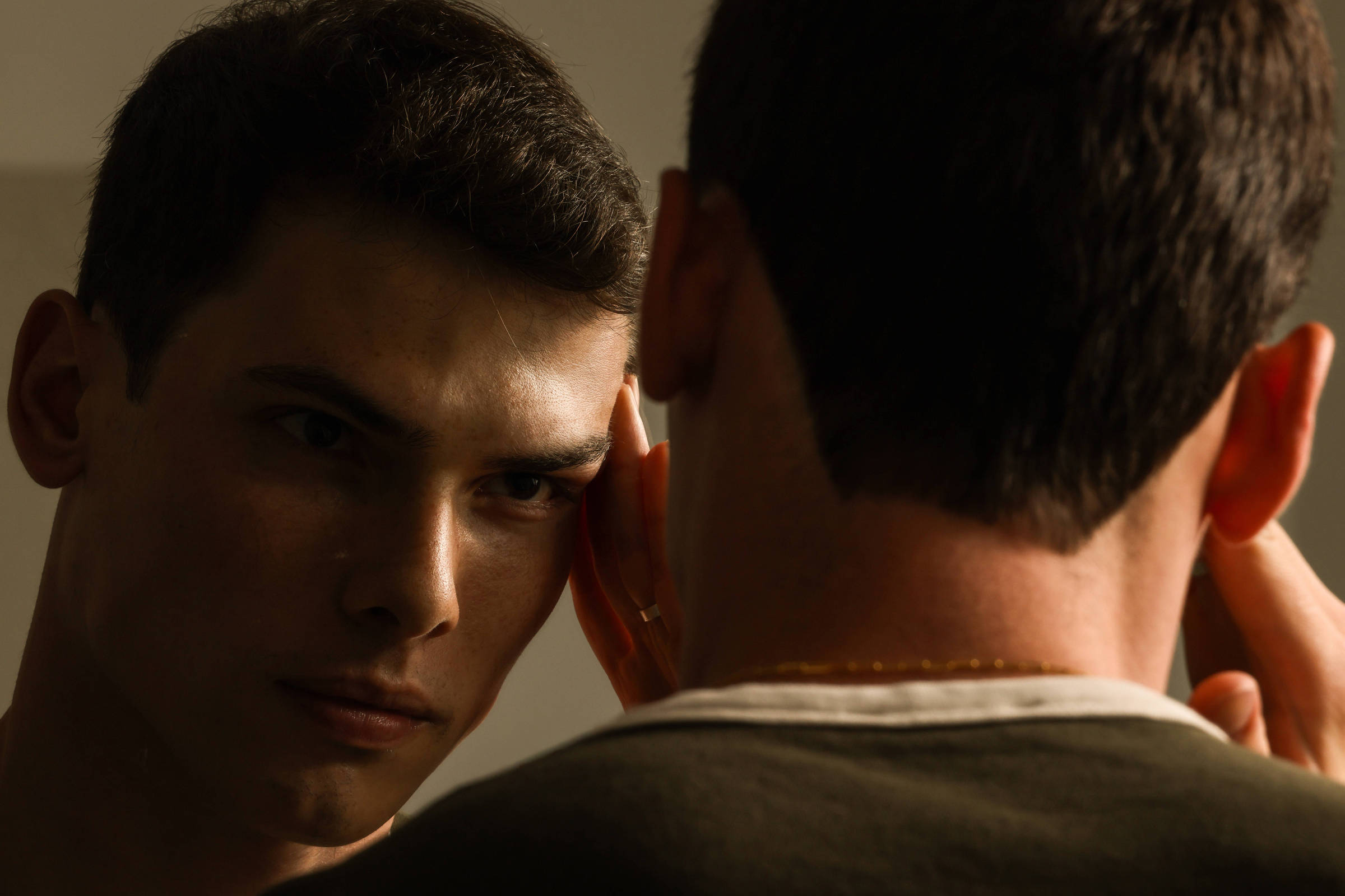 Homem de cabelo curto e escuro observa seu reflexo no espelho com expressão séria, com a mão direita tocando o rosto próximo à orelha.