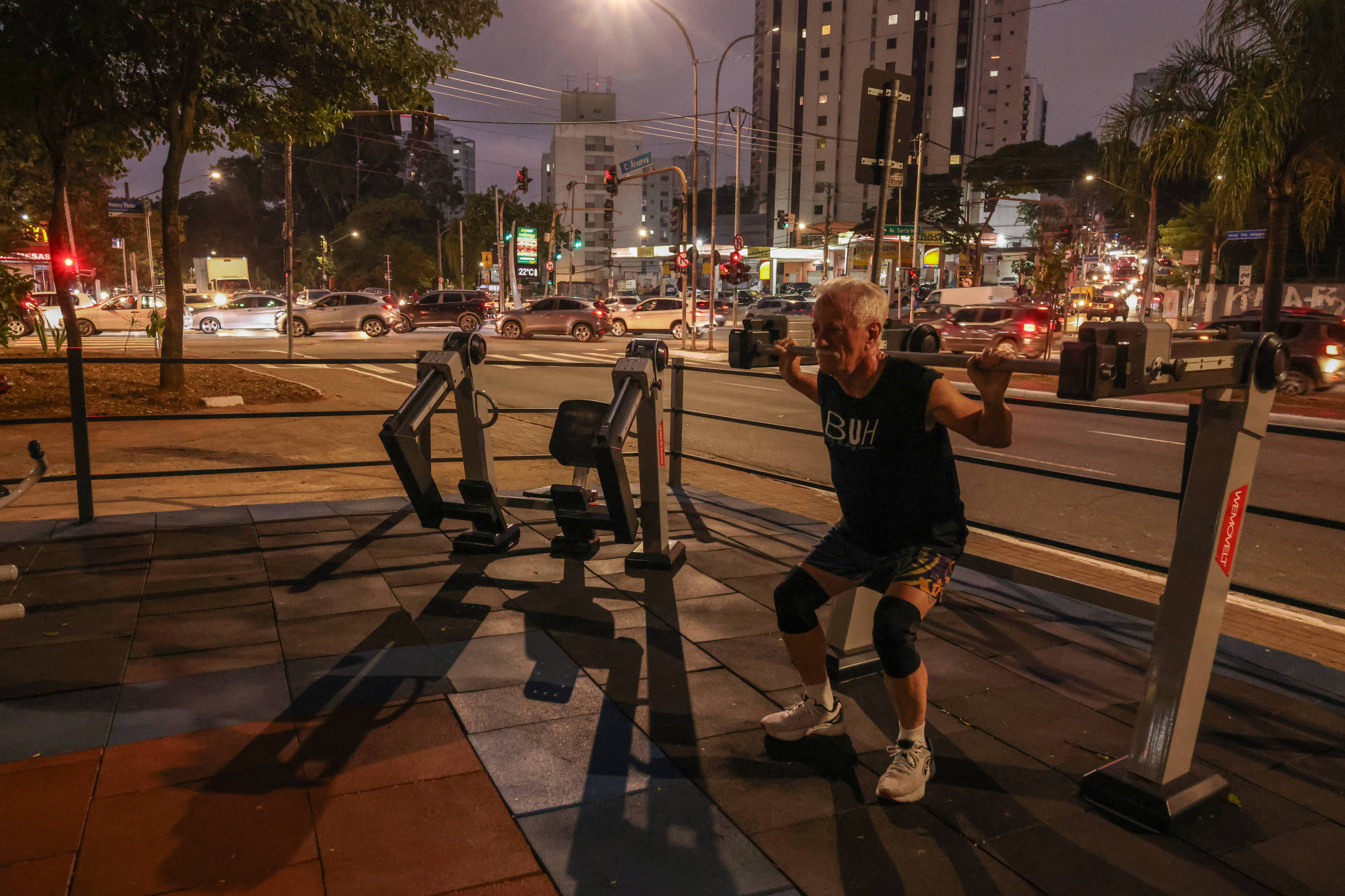 Homem idoso realiza agachamento em equipamento de academia ao ar livre próximo a avenida movimentada com carros e prédios ao fundo durante o anoitecer.