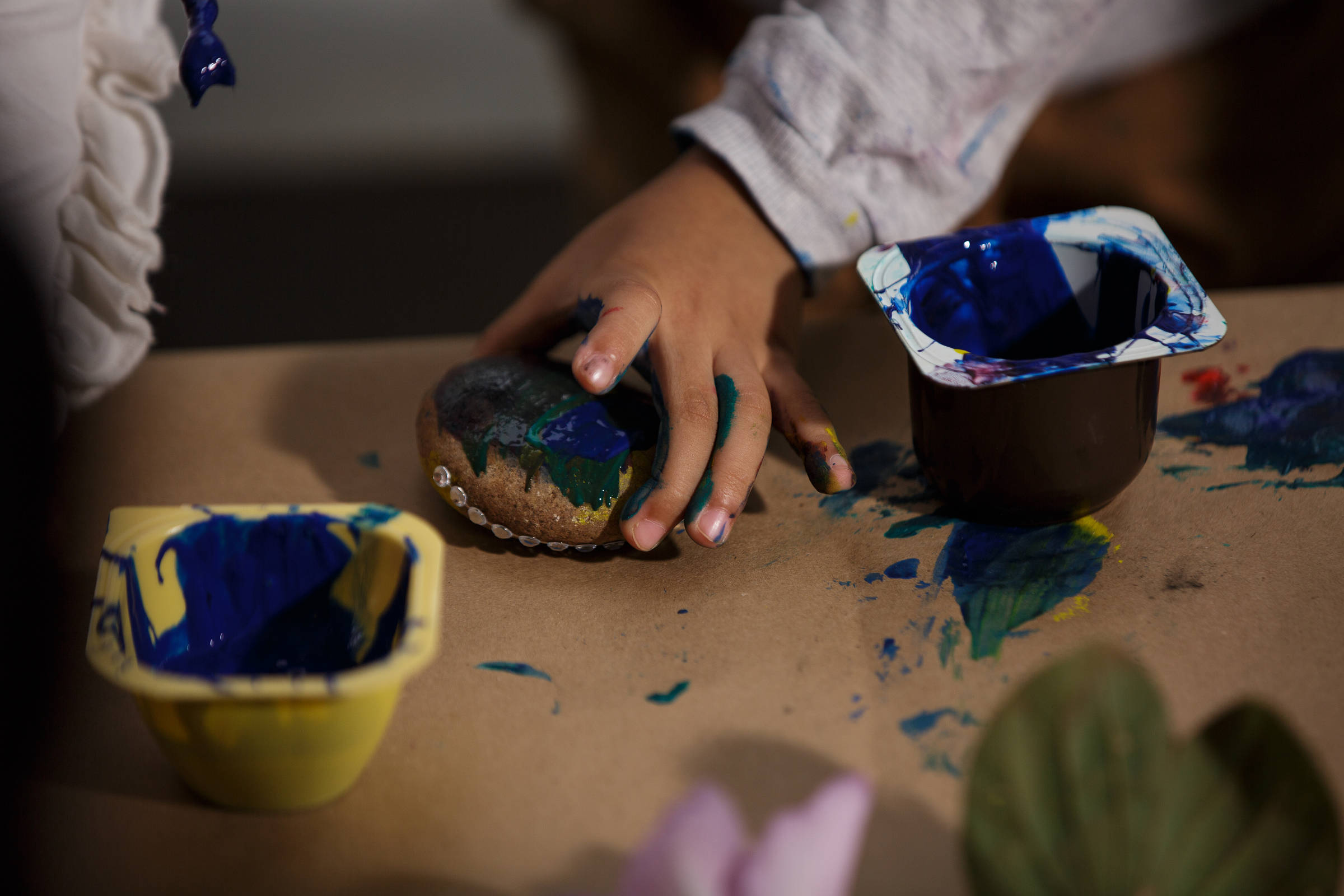 A imagem mostra uma mão de uma criança segurando uma pedra enquanto pinta com tinta azul. Ao lado, há dois recipientes com tinta, um amarelo e outro marrom, ambos com tinta azul. O fundo é uma superfície de papel marrom com manchas de tinta colorida.