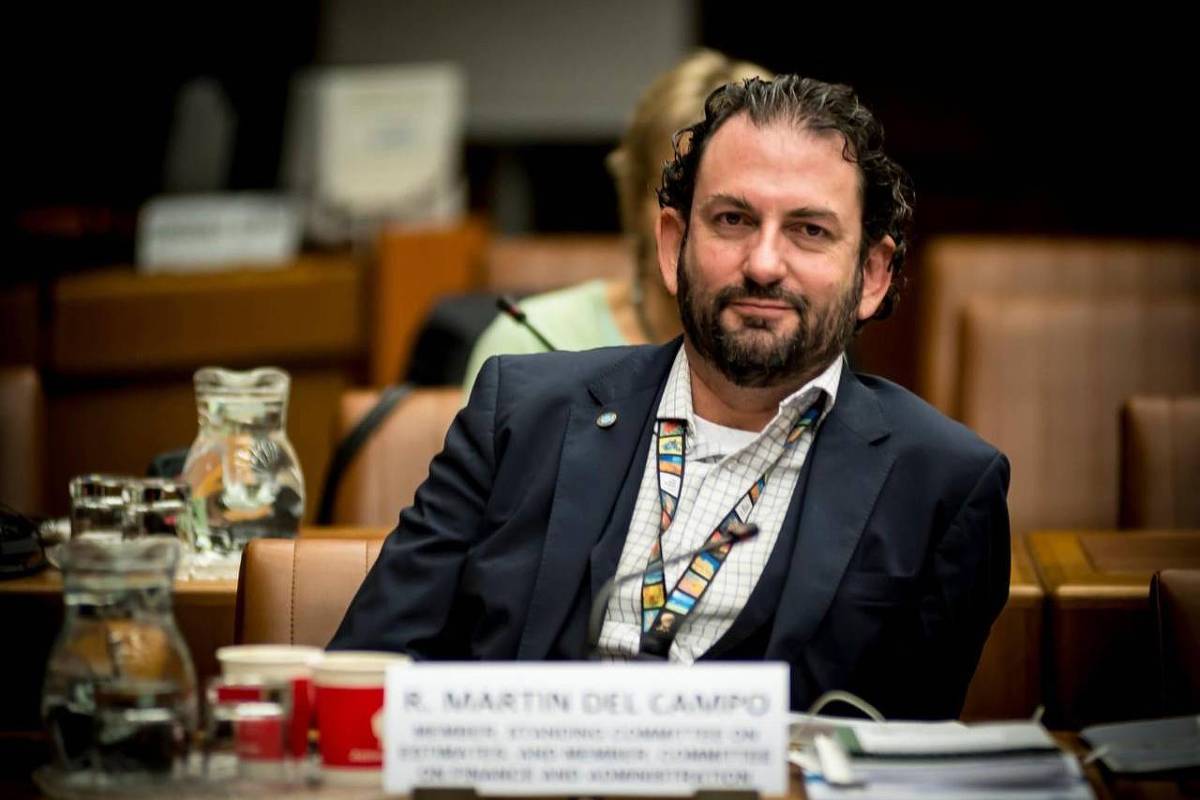 Homem de cabelos cacheados e barba, vestido com terno escuro e camisa branca, sentado em cadeira de reunião formal. À sua frente, mesa com copos, jarra de água, papéis e um crachá pendurado no pescoço. Ambiente interno com iluminação focada, outras pessoas ao fundo desfocadas.