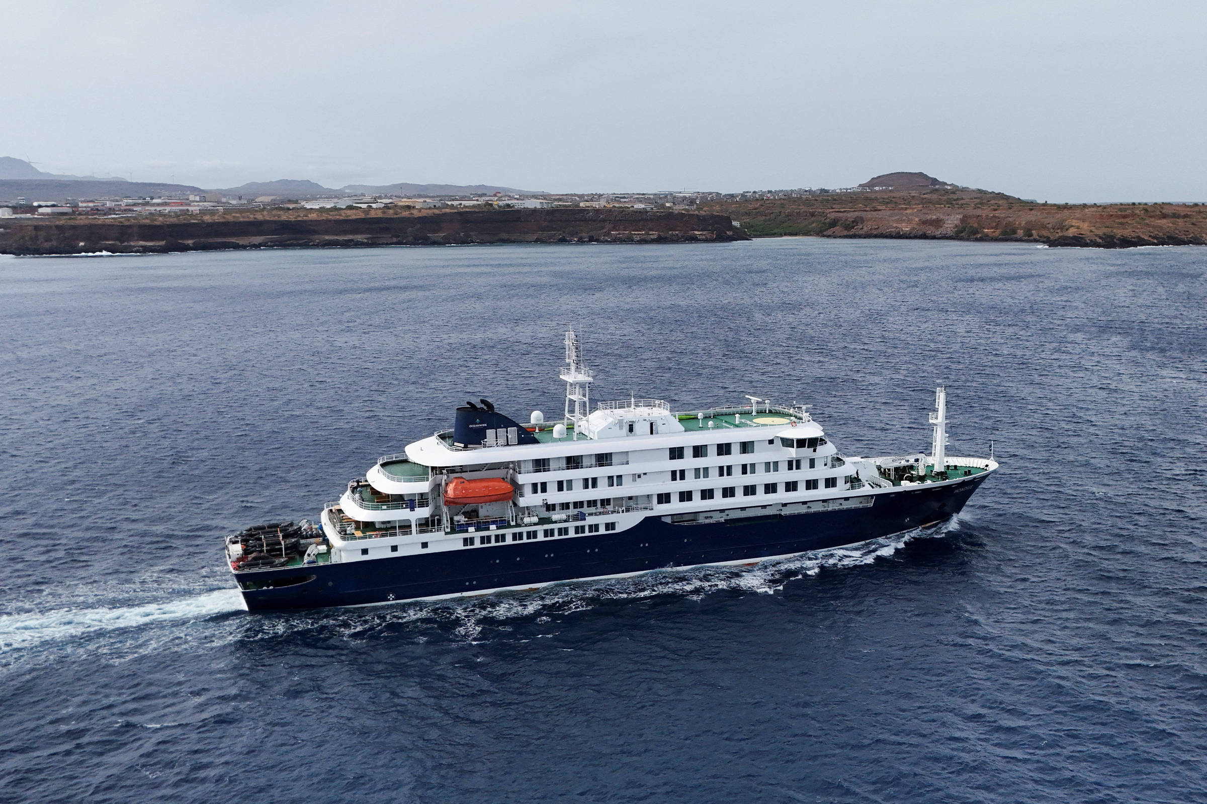 Navio de cruzeiro de médio porte com casco azul e superestrutura branca navega em mar calmo próximo a uma costa rochosa e árida sob céu nublado.