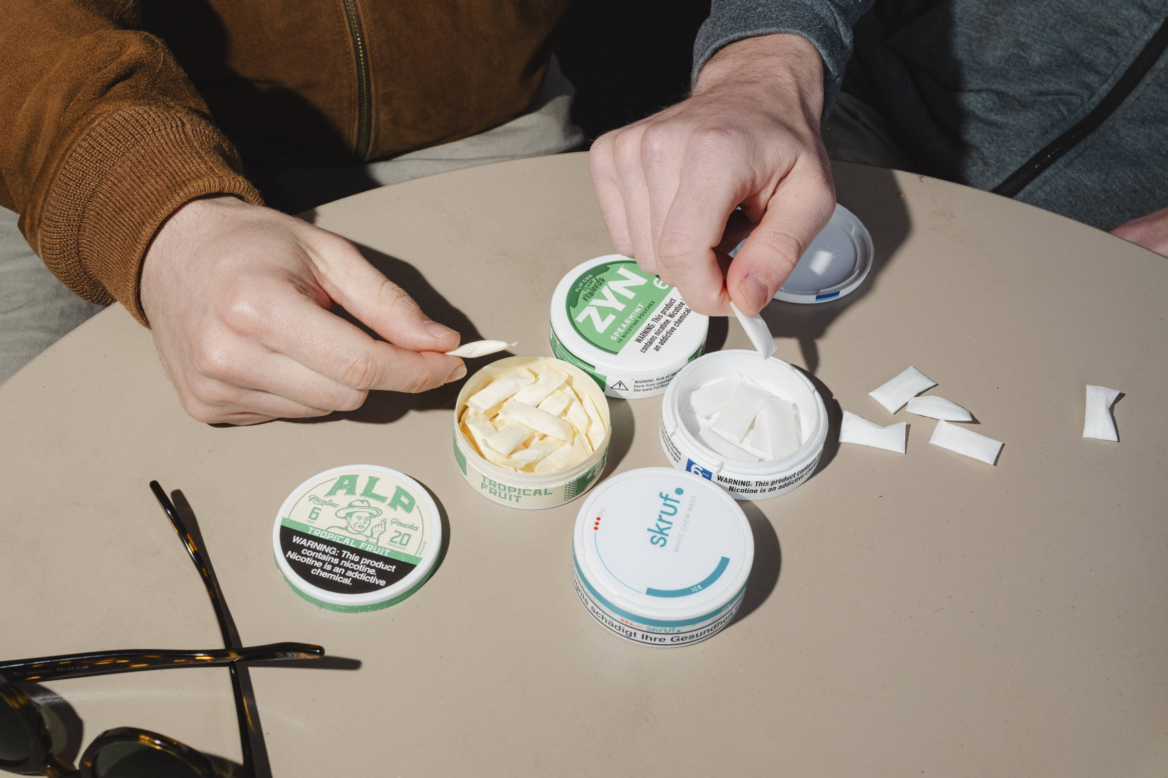 Duas pessoas com roupas casuais estão sentadas à mesa redonda clara, cada uma usando uma espátula para pegar porções de tabaco sem fumaça de recipientes abertos. Três potes de marcas diferentes estão visíveis, com tiras de tabaco ao lado. Um par de óculos escuros está apoiado na mesa.