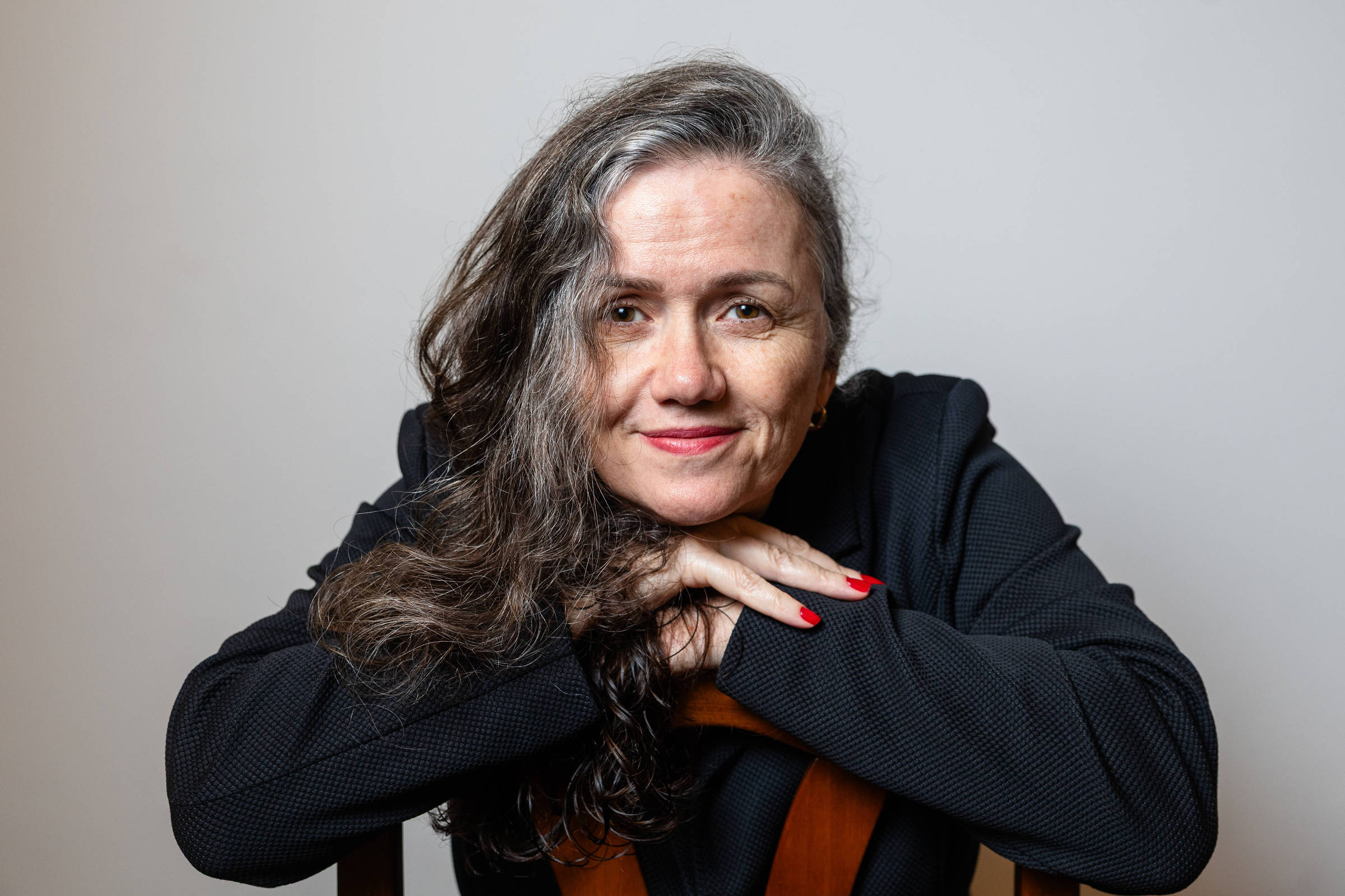 Mulher de meia-idade com cabelo longo e grisalho apoiada com os braços sobre o encosto de uma cadeira, vestindo blazer preto, com unhas pintadas de vermelho, em fundo neutro cinza.