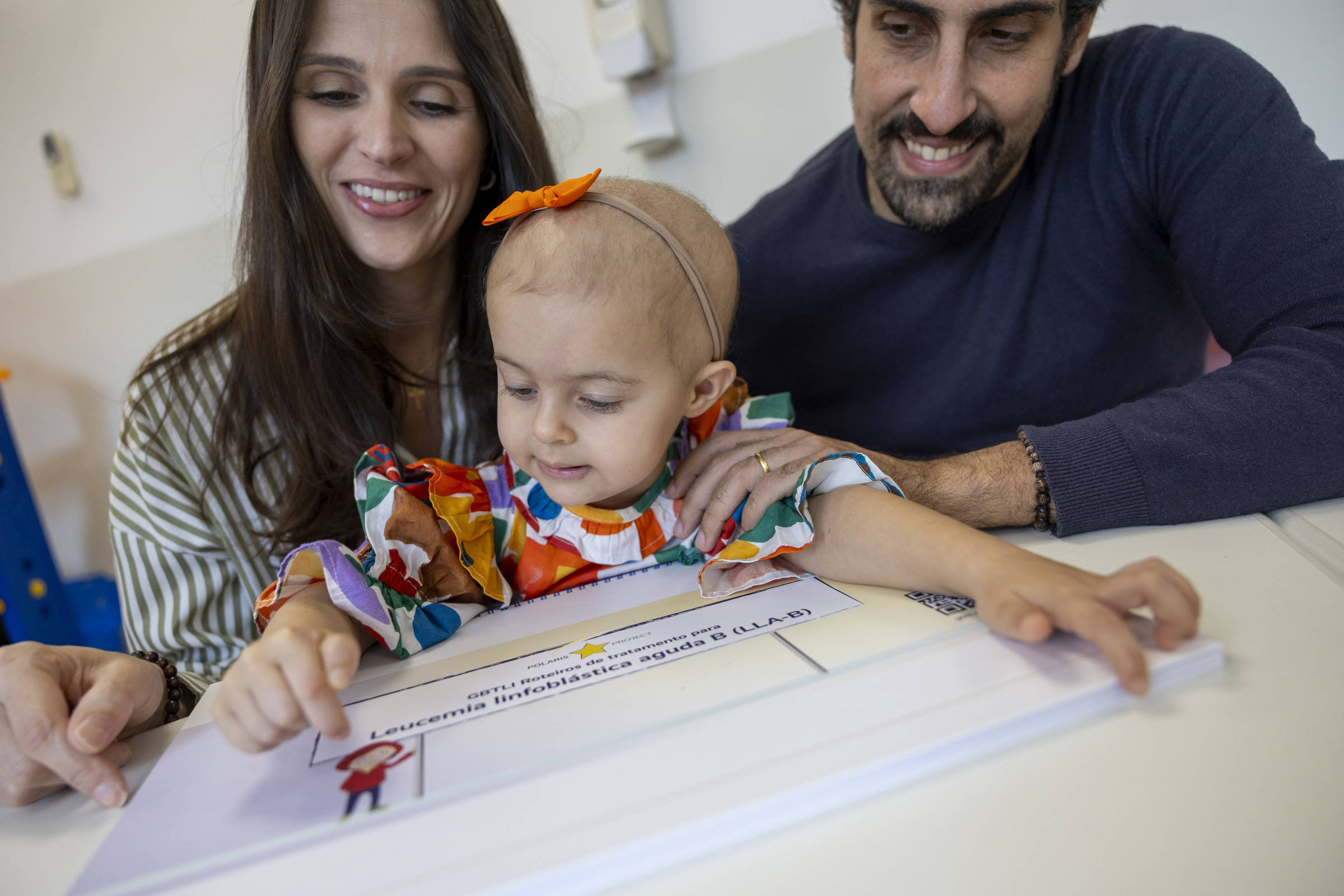 Criança pequena com faixa laranja na cabeça e vestido colorido toca material educativo sobre mesa branca, acompanhada por mulher e homem sorridentes ao fundo.