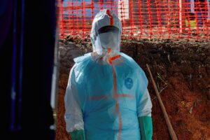Pessoa vestindo traje completo de proteção individual azul claro com detalhes laranja, máscara facial e viseira, em área externa com solo exposto e cerca de proteção laranja ao fundo.