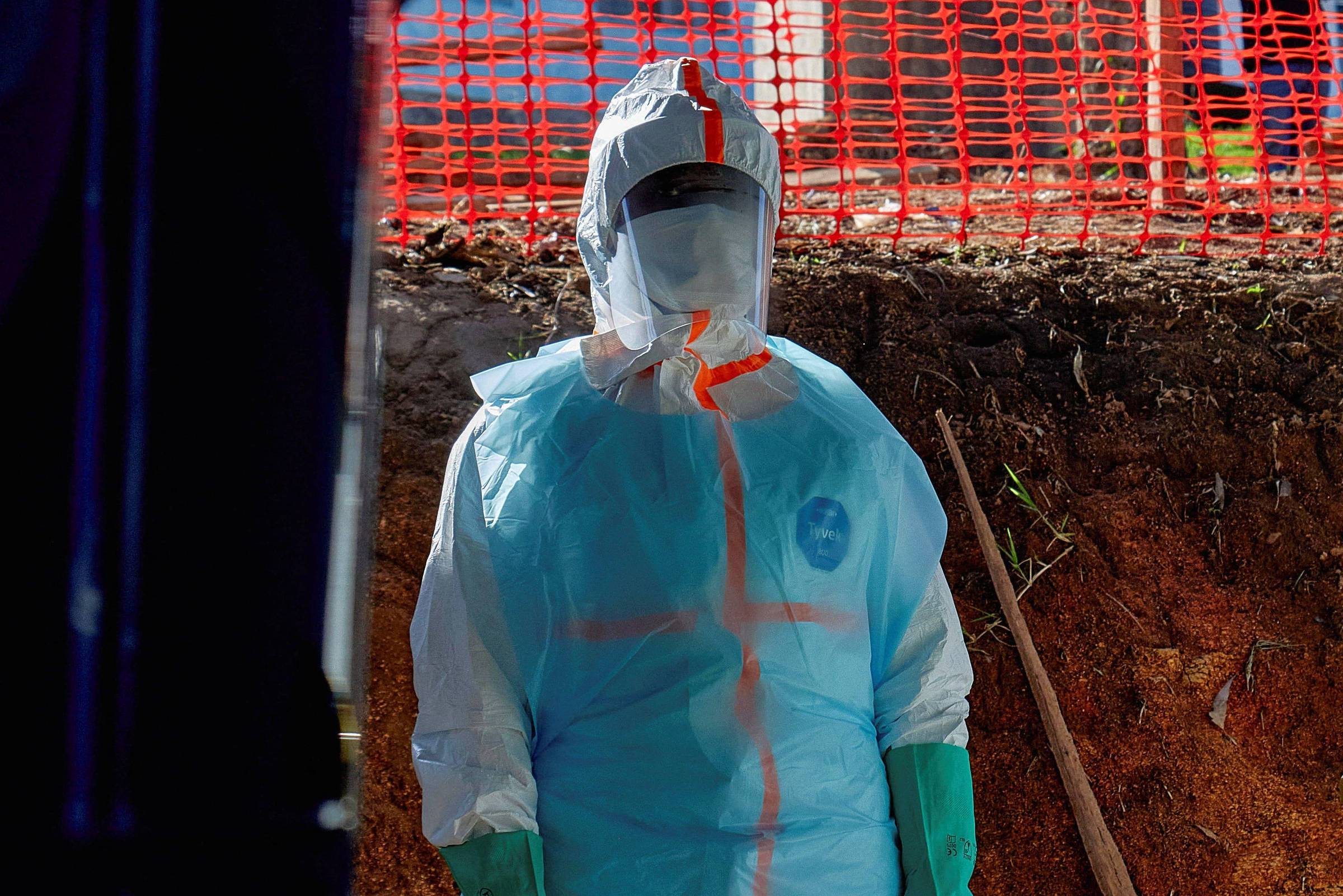 Pessoa vestindo traje completo de proteção individual azul claro com detalhes laranja, máscara facial e viseira, em área externa com solo exposto e cerca de proteção laranja ao fundo.