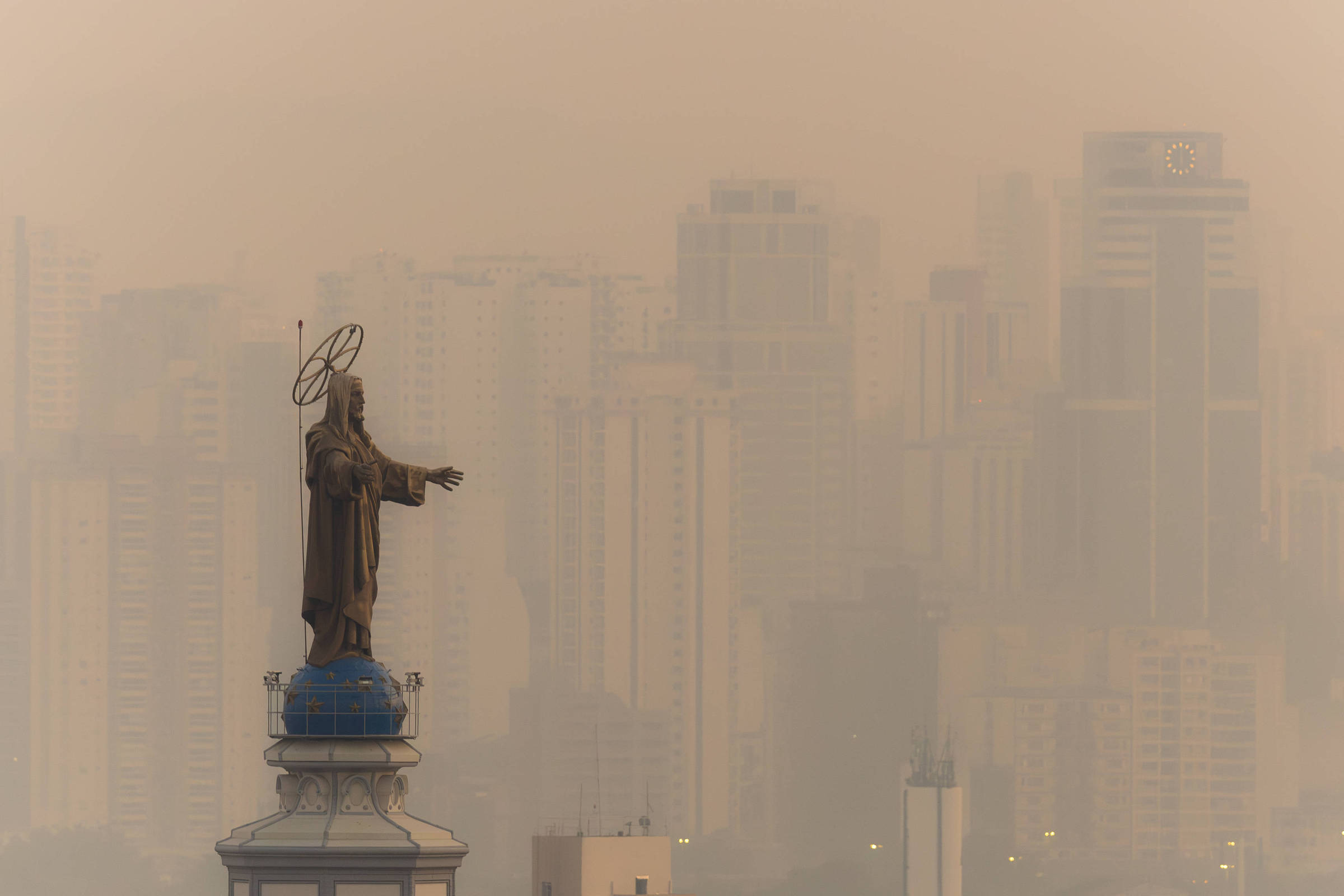 Imagem mostra estátua sobre torre de igreja com prédios ofuscados pela fumaça ao fundo