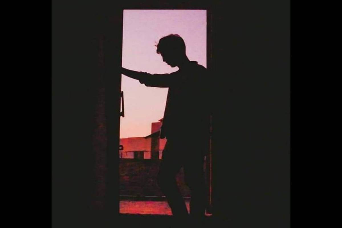 A imagem mostra a silhueta de uma pessoa em pé em uma porta, com um fundo de céu em tons de rosa e laranja, sugerindo um entardecer. A pessoa parece estar segurando algo com uma das mãos, enquanto a outra mão está apoiada na porta.