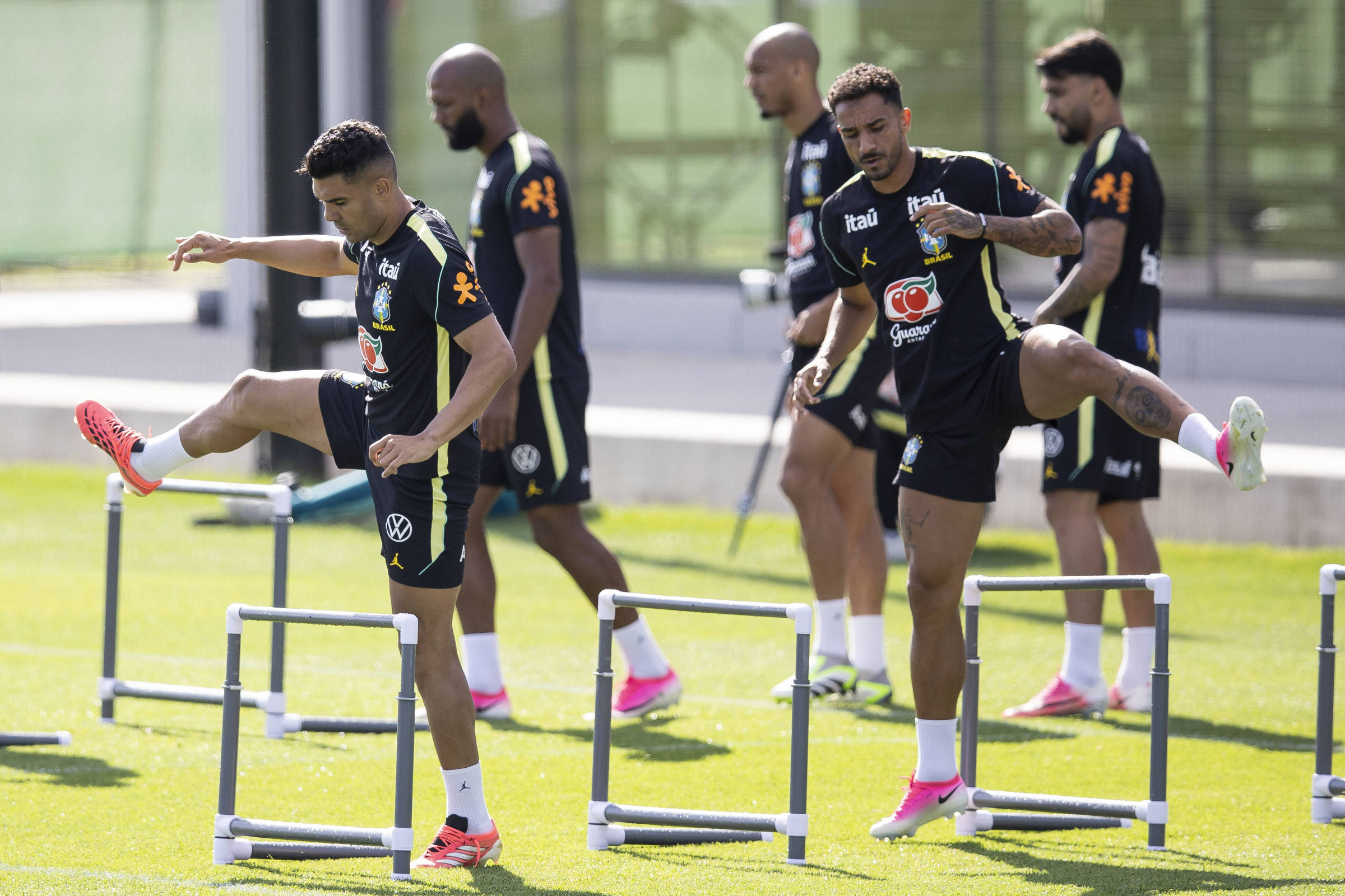 Cinco jogadores de futebol em campo gramado realizam exercícios de aquecimento pulando barreiras baixas. Dois jogadores à frente levantam as pernas sobre as barreiras, enquanto os outros três caminham ao fundo. Todos vestem uniformes pretos com detalhes amarelos e tênis esportivos coloridos.
