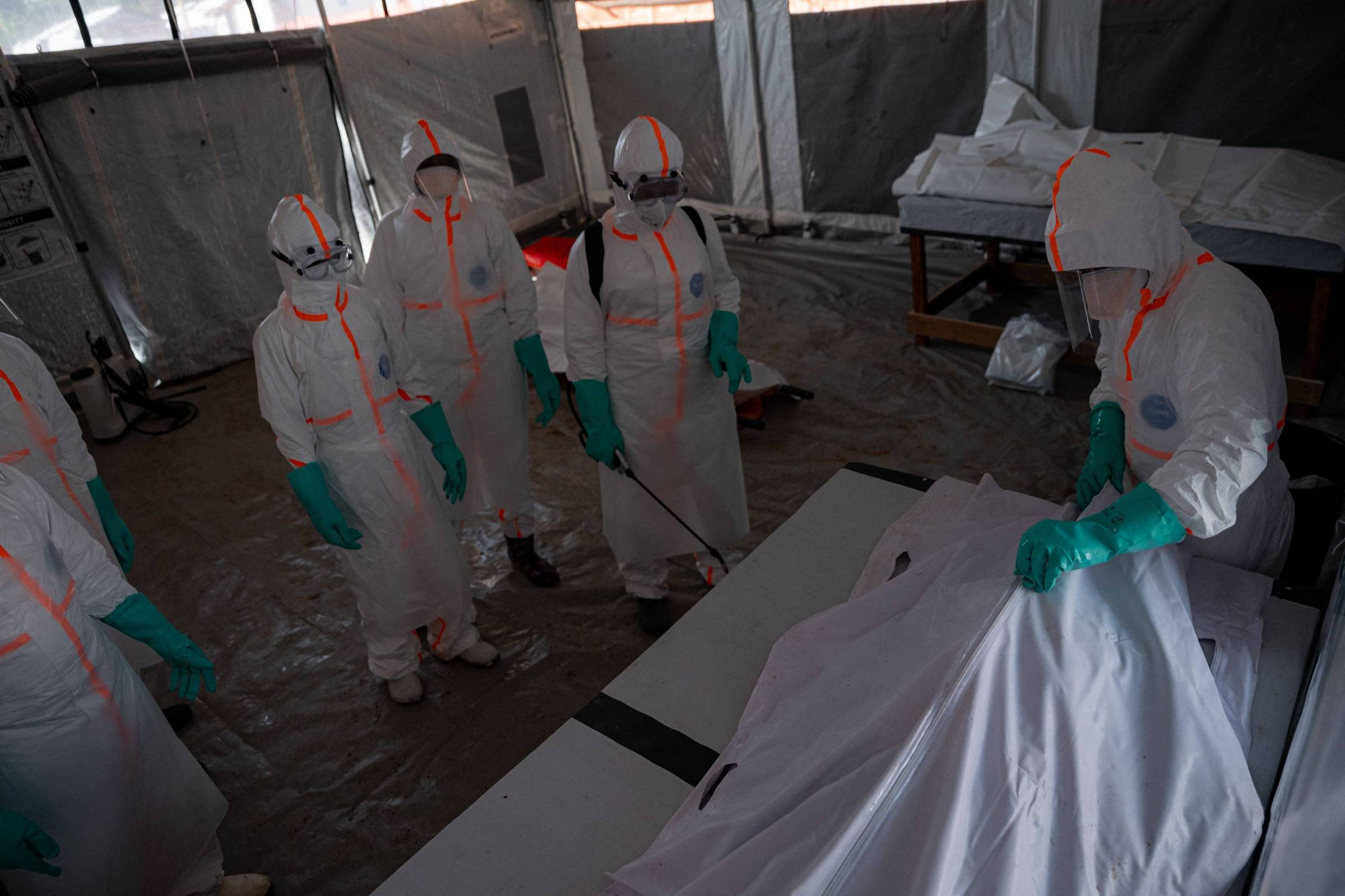 Cinco pessoas vestindo trajes de proteção branca com detalhes laranja e luvas verdes estão em uma tenda. Uma delas segura um corpo coberto por um lençol branco sobre uma maca, enquanto as outras observam ao redor.