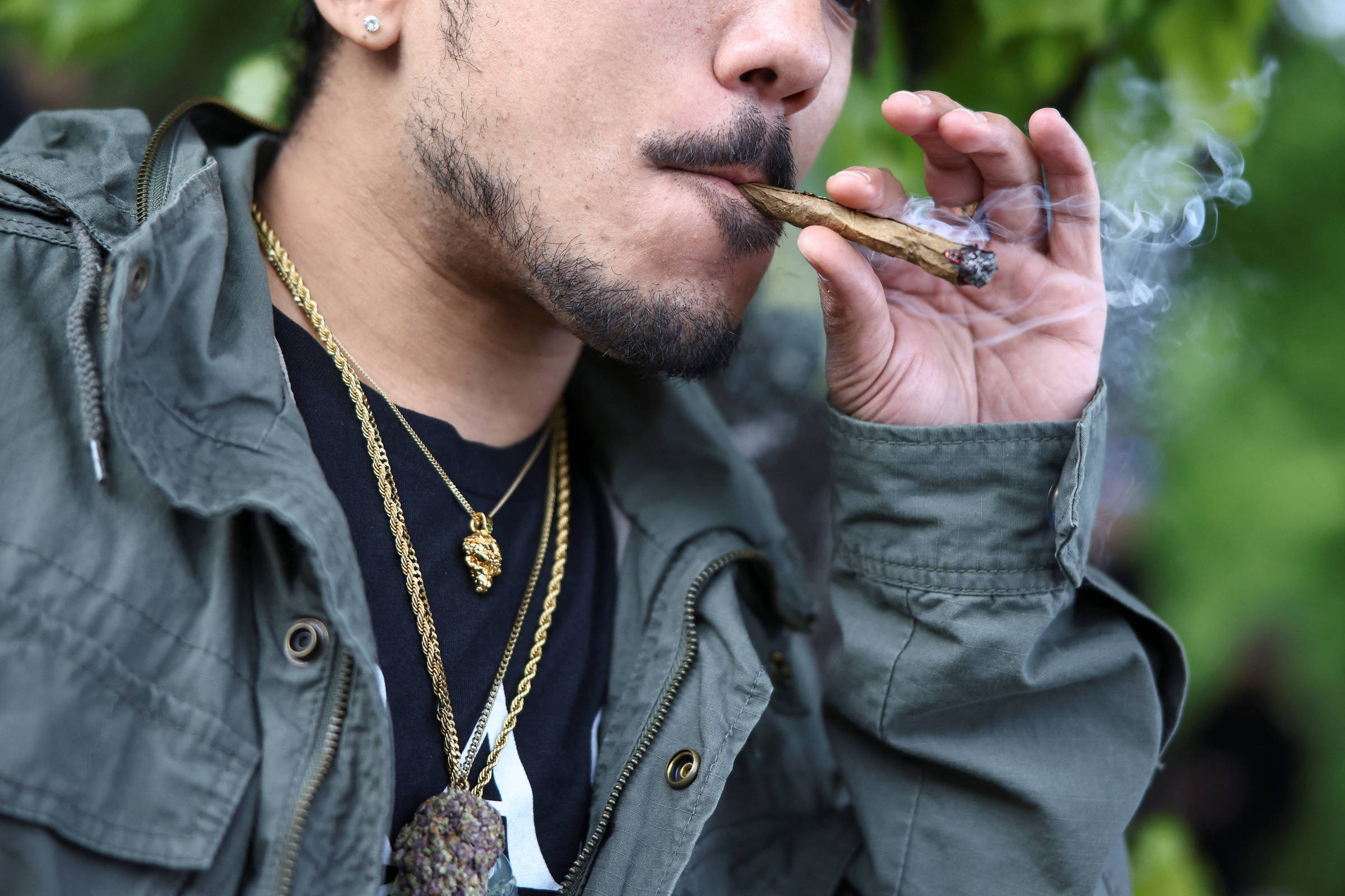 Homem com jaqueta verde e colares dourados segura e fuma cigarro enrolado, com fumaça visível. Fundo desfocado com vegetação.