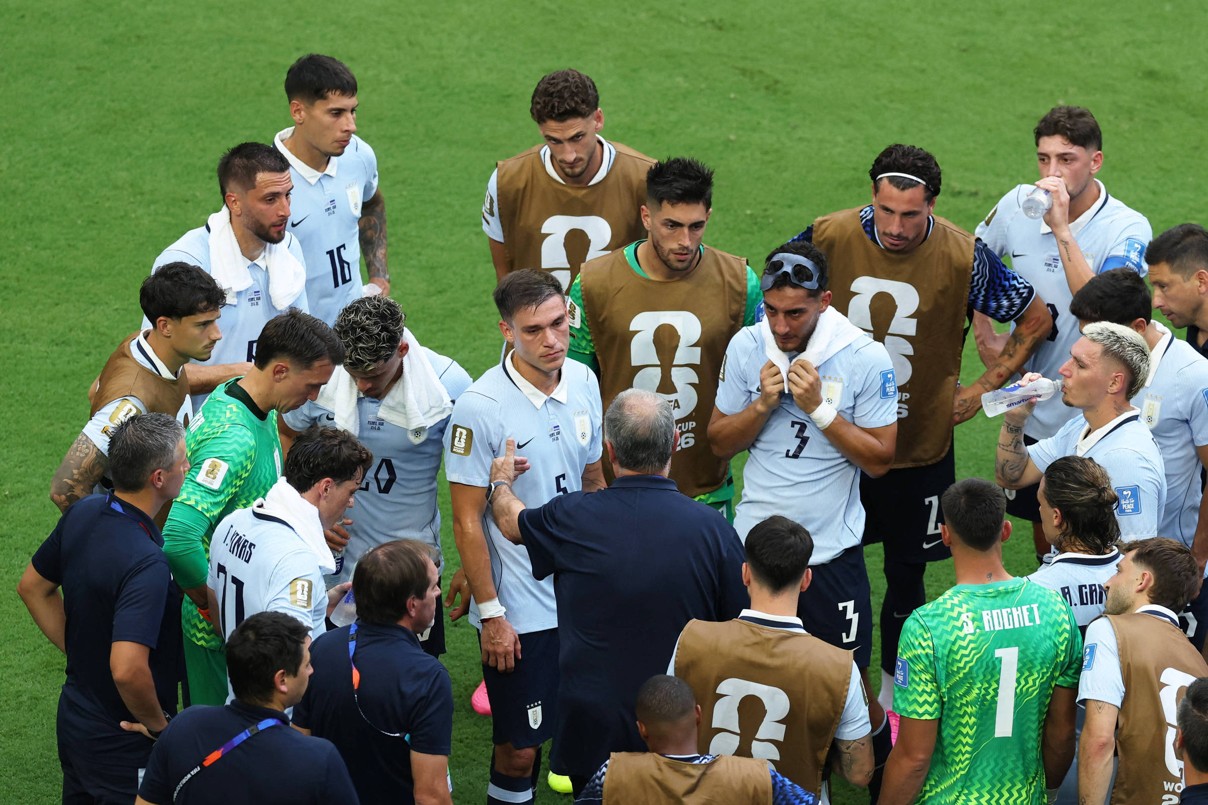Jogadores da seleção uruguaia de futebol reunidos em círculo no campo, ouvindo orientações do técnico. Alguns atletas vestem coletes marrons, outros o uniforme branco com detalhes azuis, em gramado verde.