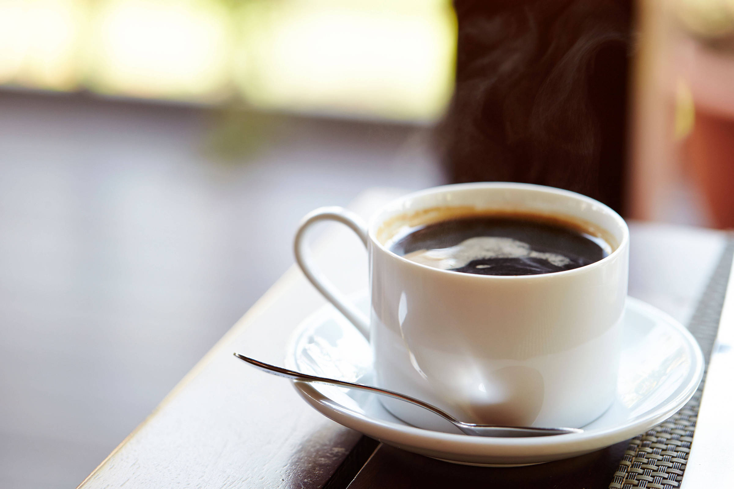 Xícara branca cheia de café preto com vapor saindo, apoiada em pires branco sobre mesa de madeira clara. Colher de metal repousa no pires ao lado da xícara.