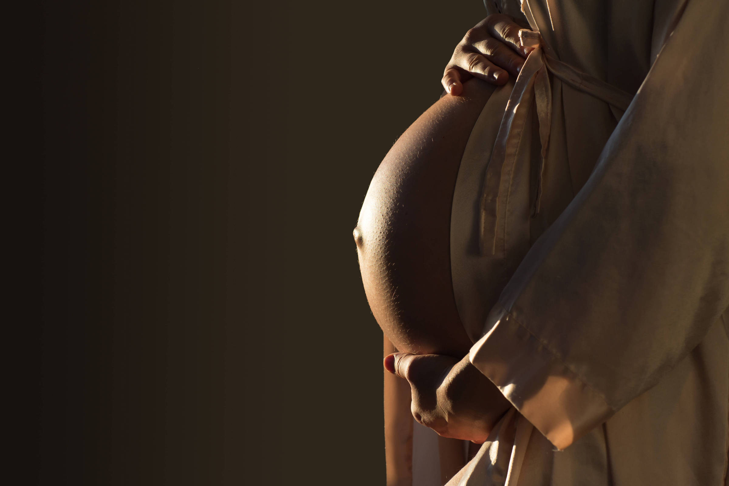 A imagem mostra uma mulher grávida segurando sua barriga com as duas mãos. Ela está vestindo uma roupa clara e a iluminação destaca a forma arredondada da barriga. O fundo é escuro, criando um contraste com a figura da mulher.