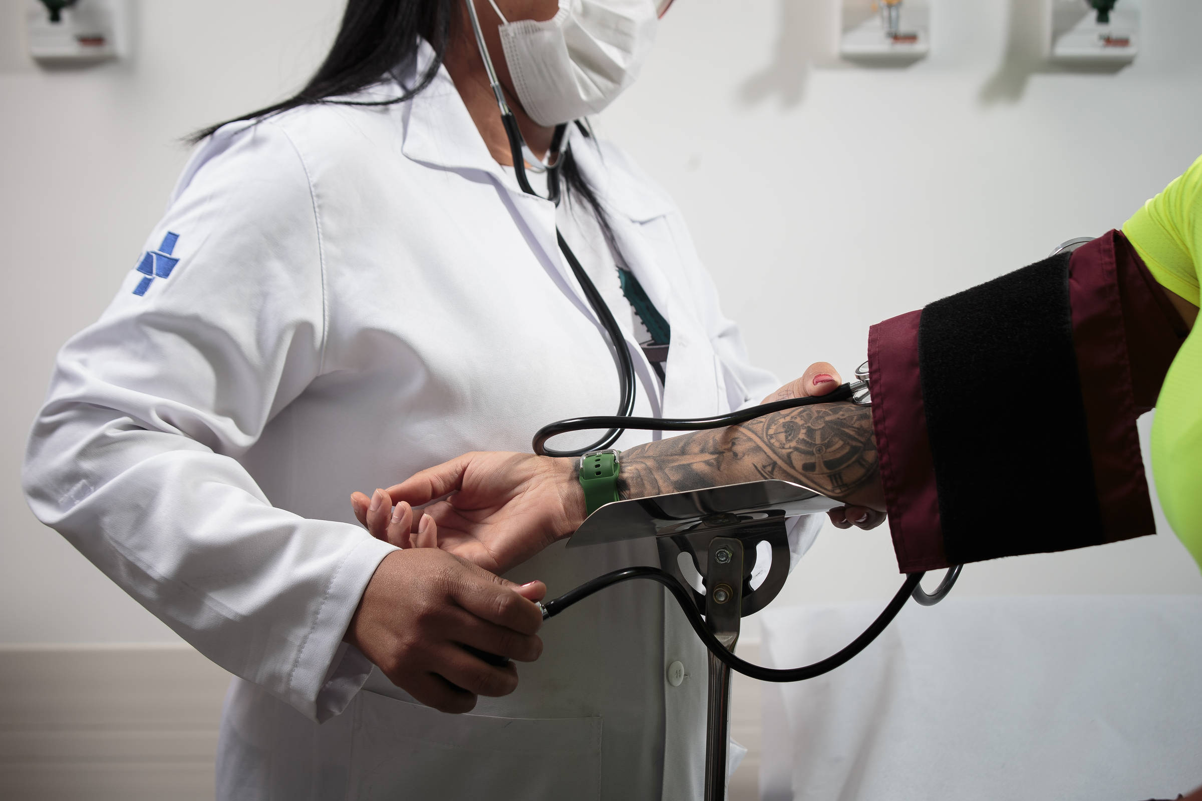 Uma profissional de saúde, vestindo um jaleco branco e máscara, está medindo a pressão arterial de um paciente. O paciente tem um braço tatuado e está com um manguito de pressão no braço. O ambiente parece ser uma sala de consulta médica, com equipamentos de saúde ao fundo.