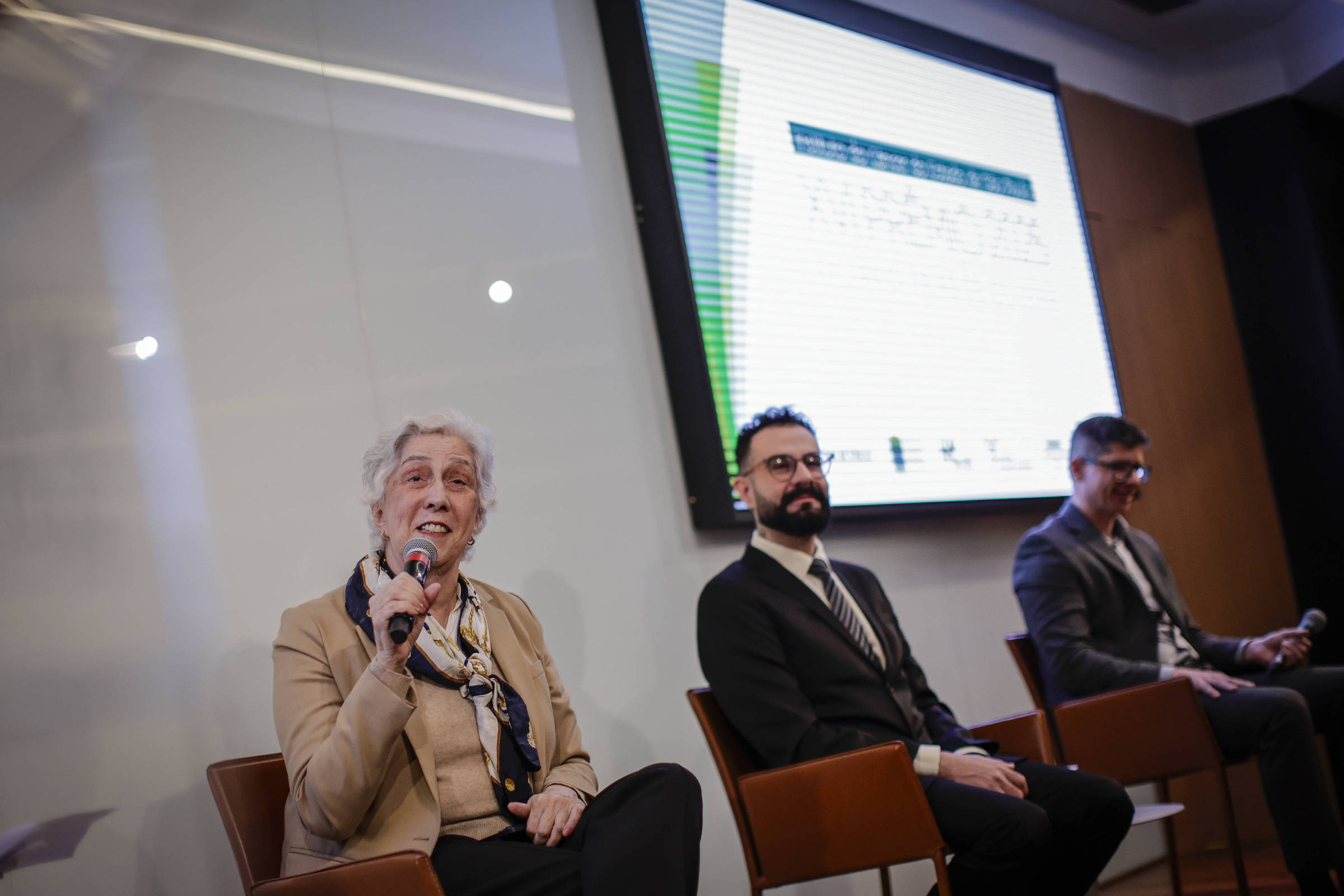 A imagem mostra um painel de discussão com três pessoas sentadas. À esquerda, uma mulher de cabelo grisalho, usando um blazer claro e segurando um microfone, parece estar falando. No centro, um homem com barba e óculos, vestido com um terno escuro e gravata, escuta atentamente. À direita, outro homem também de terno, está sentado e observa. Ao fundo, uma tela exibe informações, mas o conteúdo não é legível.
