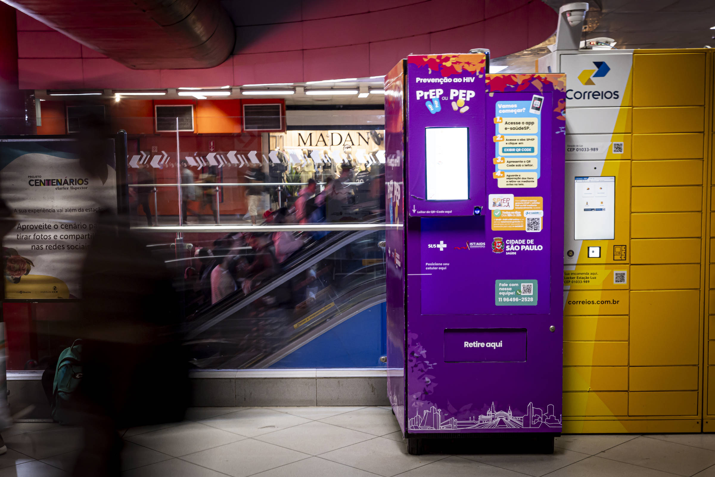 A imagem mostra duas máquinas em uma estação de metrô. À esquerda, há uma máquina roxa com o nome
