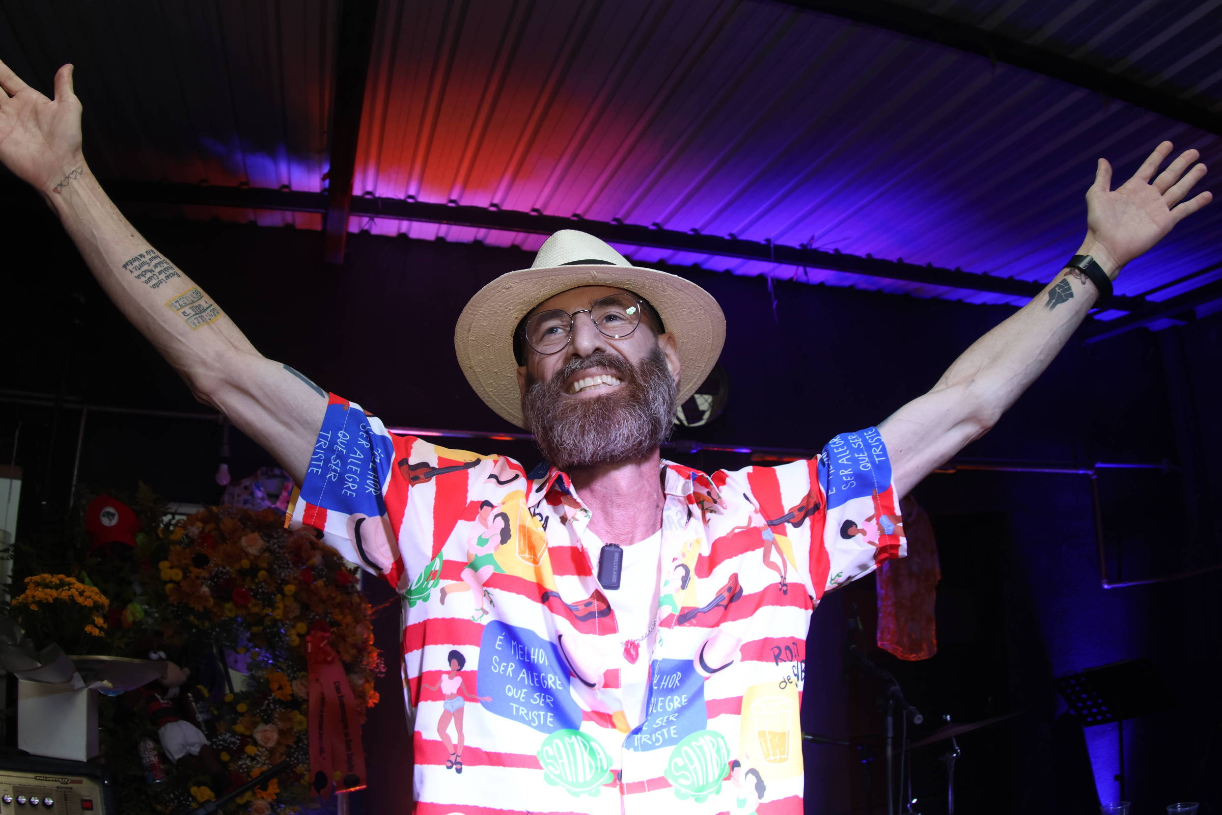 Homem de barba e óculos usa chapéu branco e camisa colorida com estampas diversas, com os braços abertos em gesto amplo. Ele está em ambiente interno com iluminação roxa e vermelha no teto metálico.