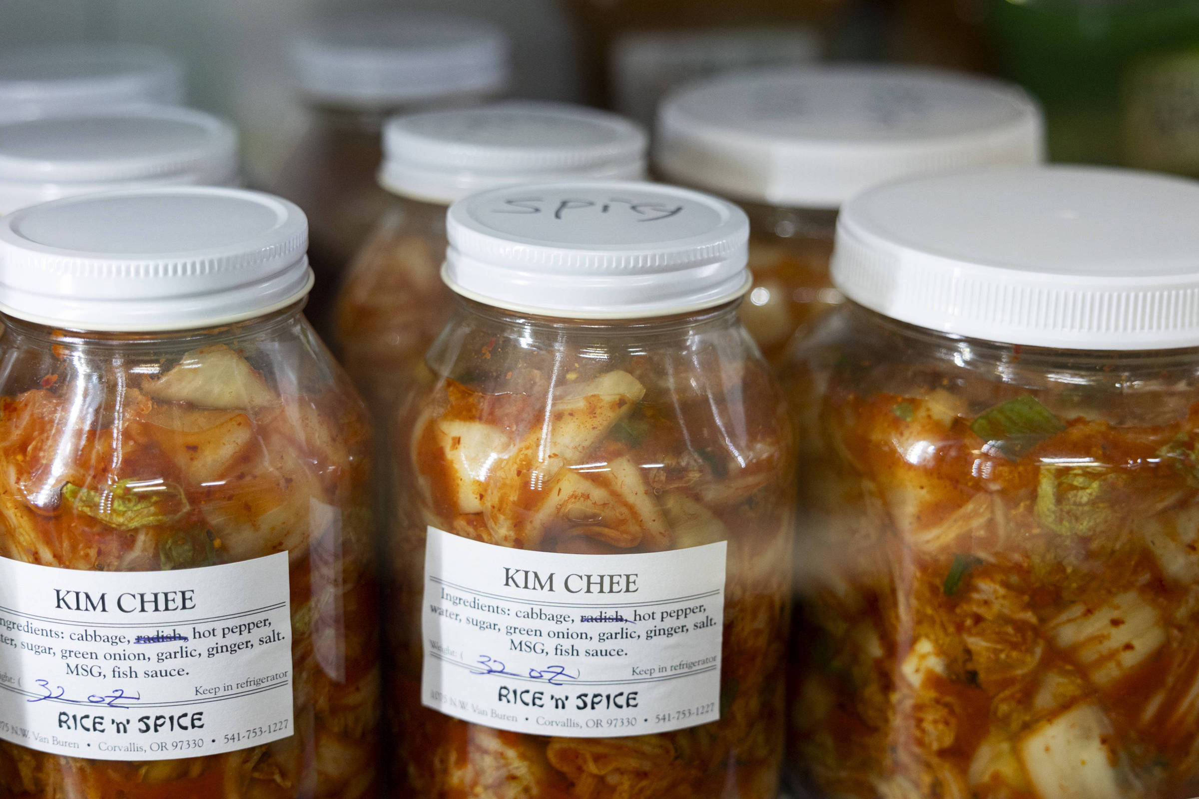 Vários potes transparentes com tampa branca contendo kimchee, um prato fermentado de vegetais. As etiquetas indicam ingredientes como repolho, rabanete, pimenta, alho e gengibre, e mencionam a marca Rice