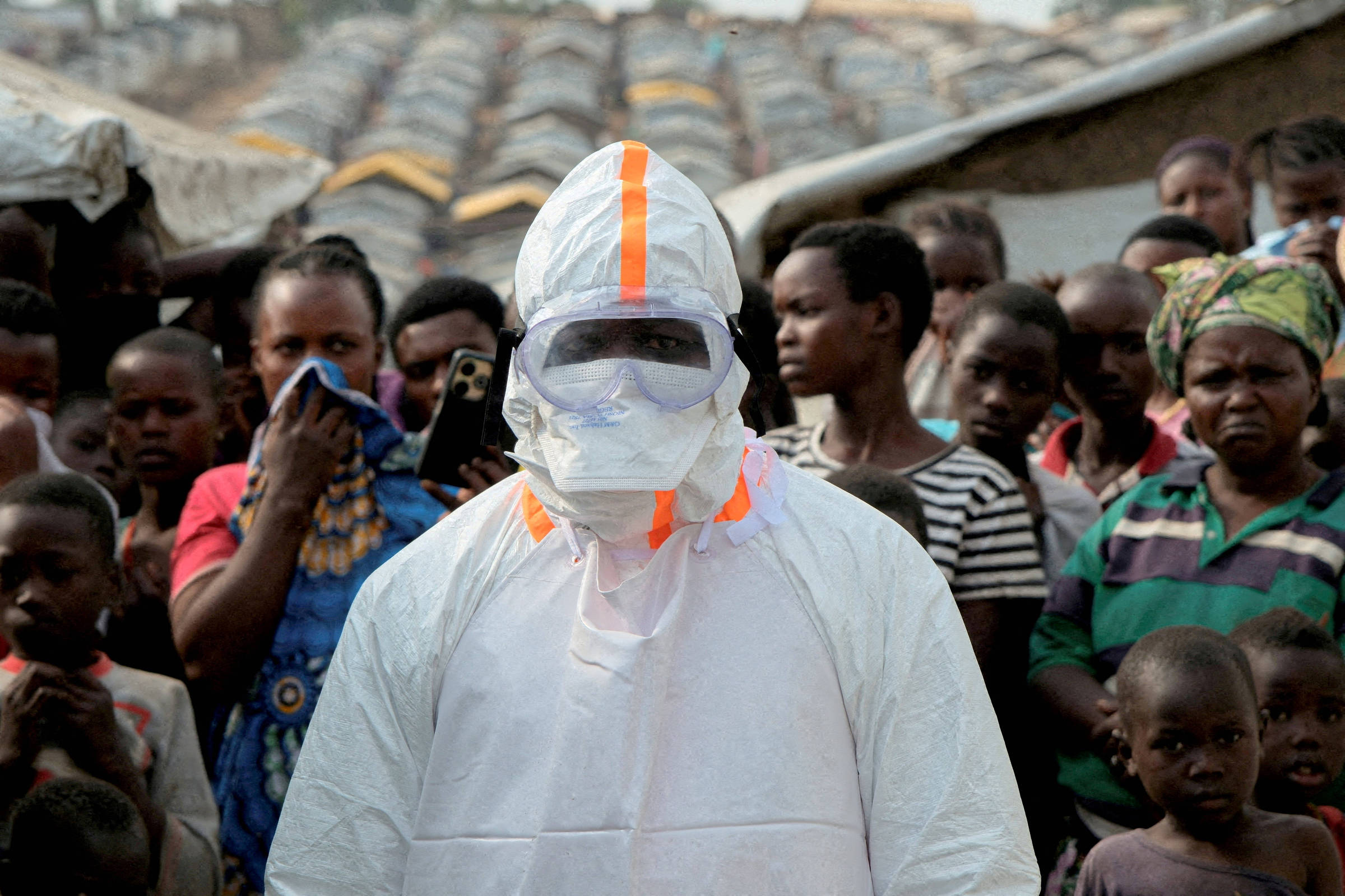 Pessoa vestindo traje branco de proteção com faixa laranja e óculos de segurança está em primeiro plano, cercada por grupo de moradores locais, incluindo mulheres e crianças, em área com casas de telhado de barro ao fundo.