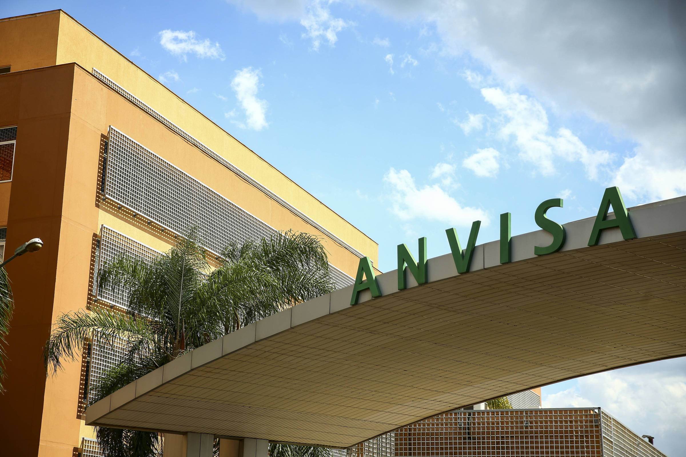 Fachada do prédio da Anvisa em dia claro com céu parcialmente nublado. Letreiro verde com as letras
