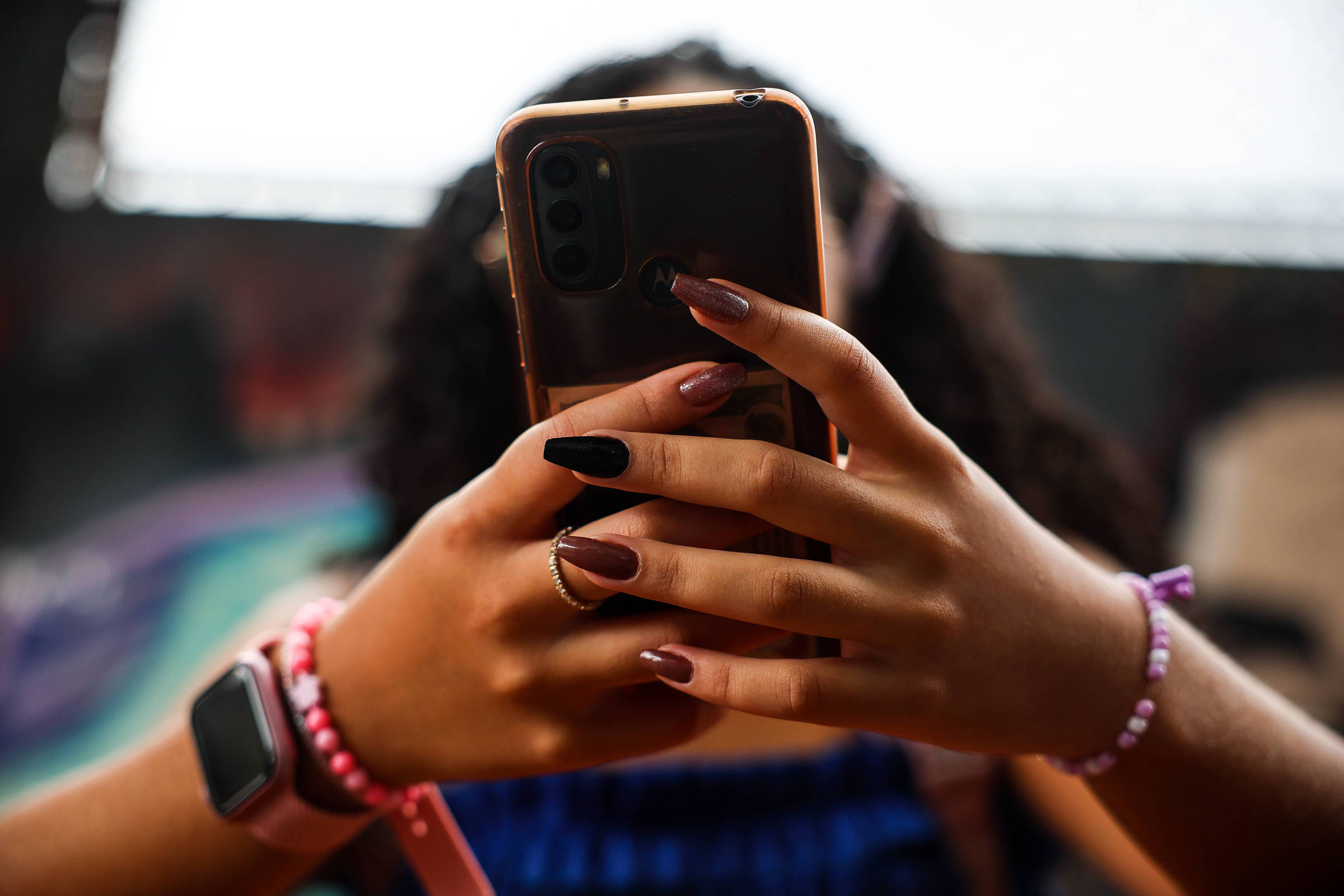 Mãos femininas seguram smartphone com capa transparente. Unhas longas pintadas em tons de marrom e preto. Pulso com relógio digital rosa e pulseiras de contas rosa e lilás. Fundo desfocado com luz natural.