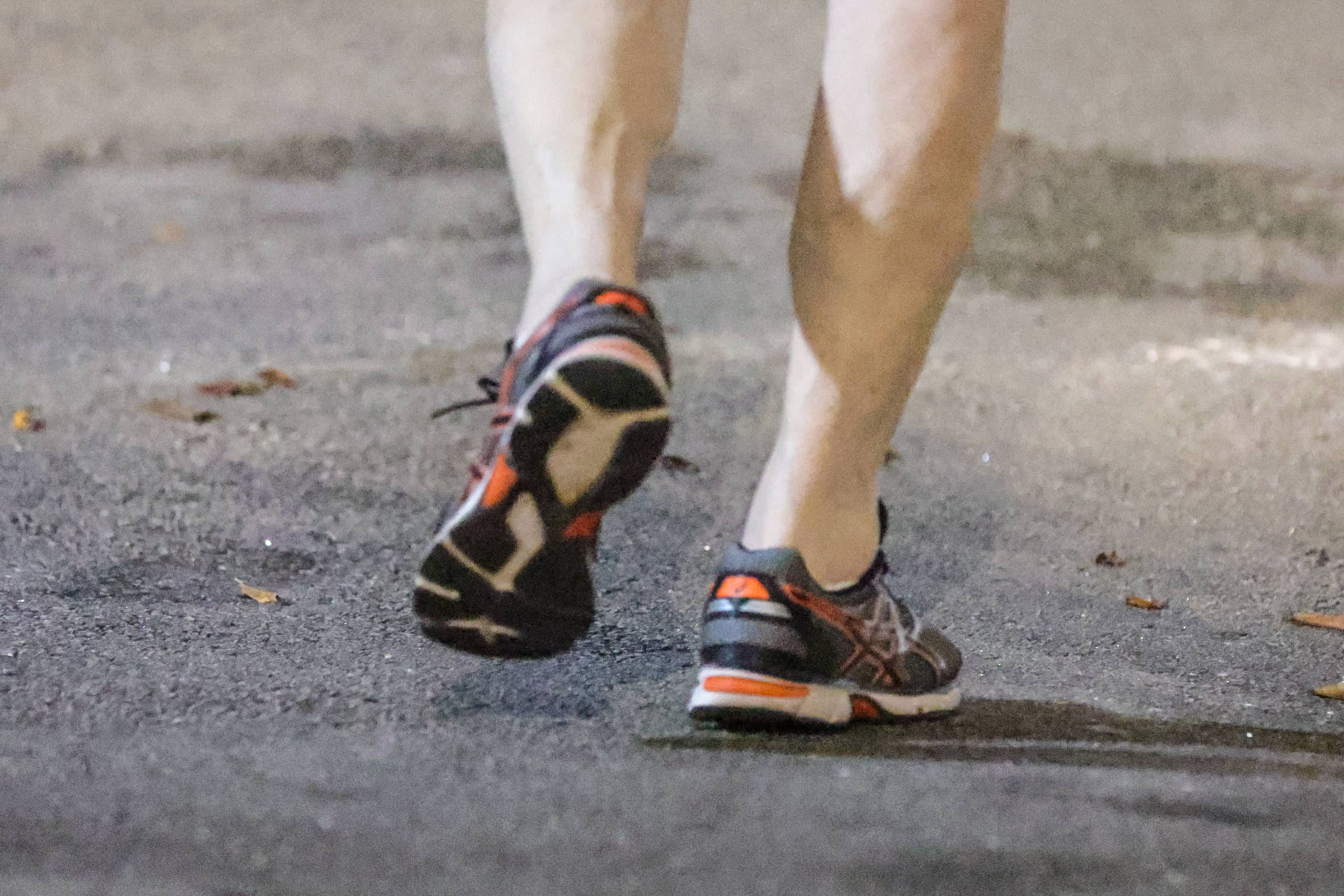 A imagem mostra as pernas de uma pessoa em movimento, com foco nos pés calçados com tênis de corrida. O chão é de asfalto e há algumas folhas secas ao redor. O tênis é predominantemente escuro com detalhes em laranja e branco.