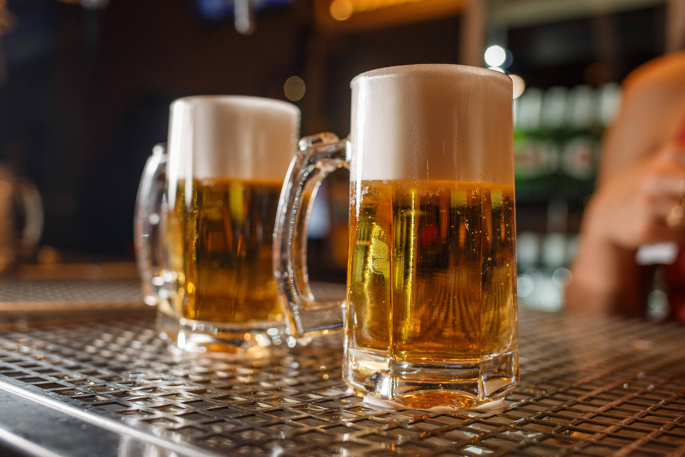 Duas canecas de cerveja com espuma no topo, colocadas sobre uma superfície metálica. A cerveja é de cor dourada e translúcida, e ao fundo, há uma leve desfocagem que sugere um ambiente de bar.