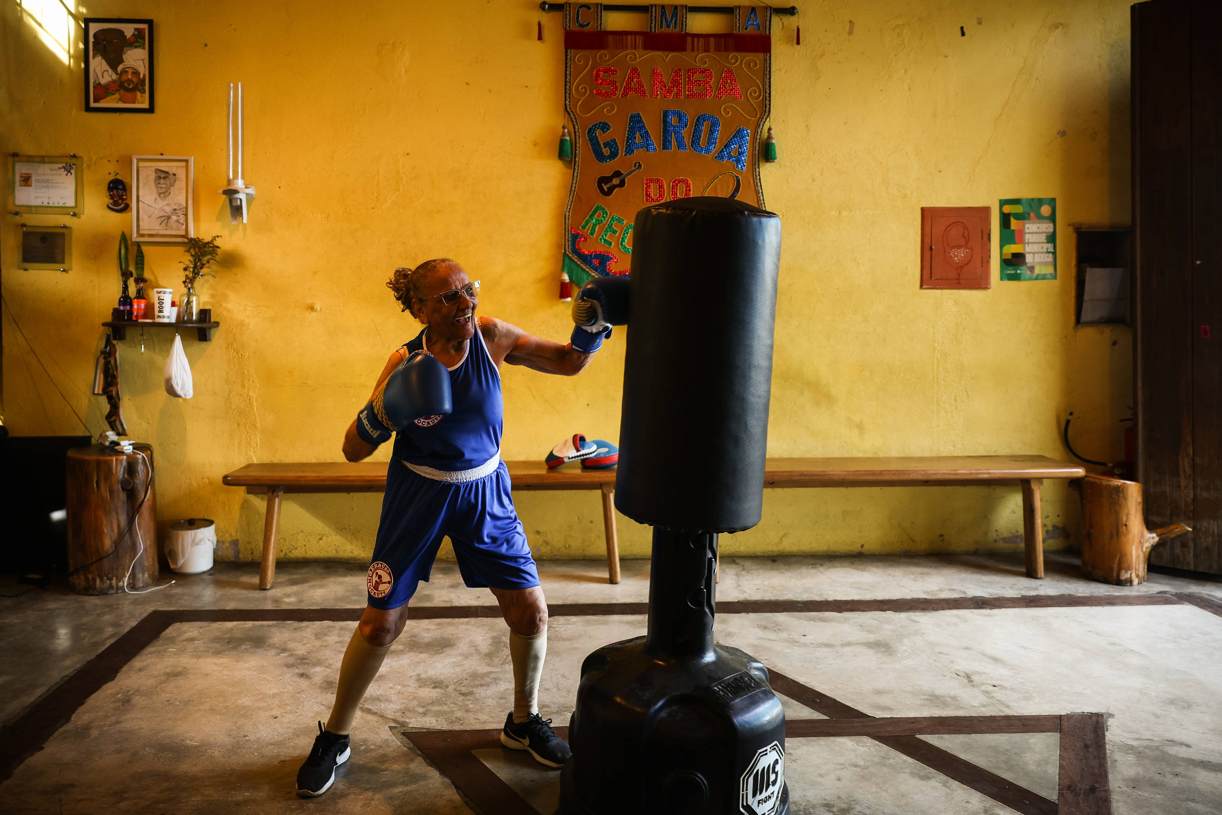 Homem idoso com roupa azul de boxe e luvas treina socos em saco de pancadas preto dentro de academia com paredes amarelas e decoração simples.